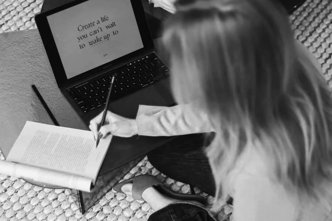 Pessoa escrevendo em um livro aberto com uma caneta, ao lado de um laptop que exibe uma frase motivacional