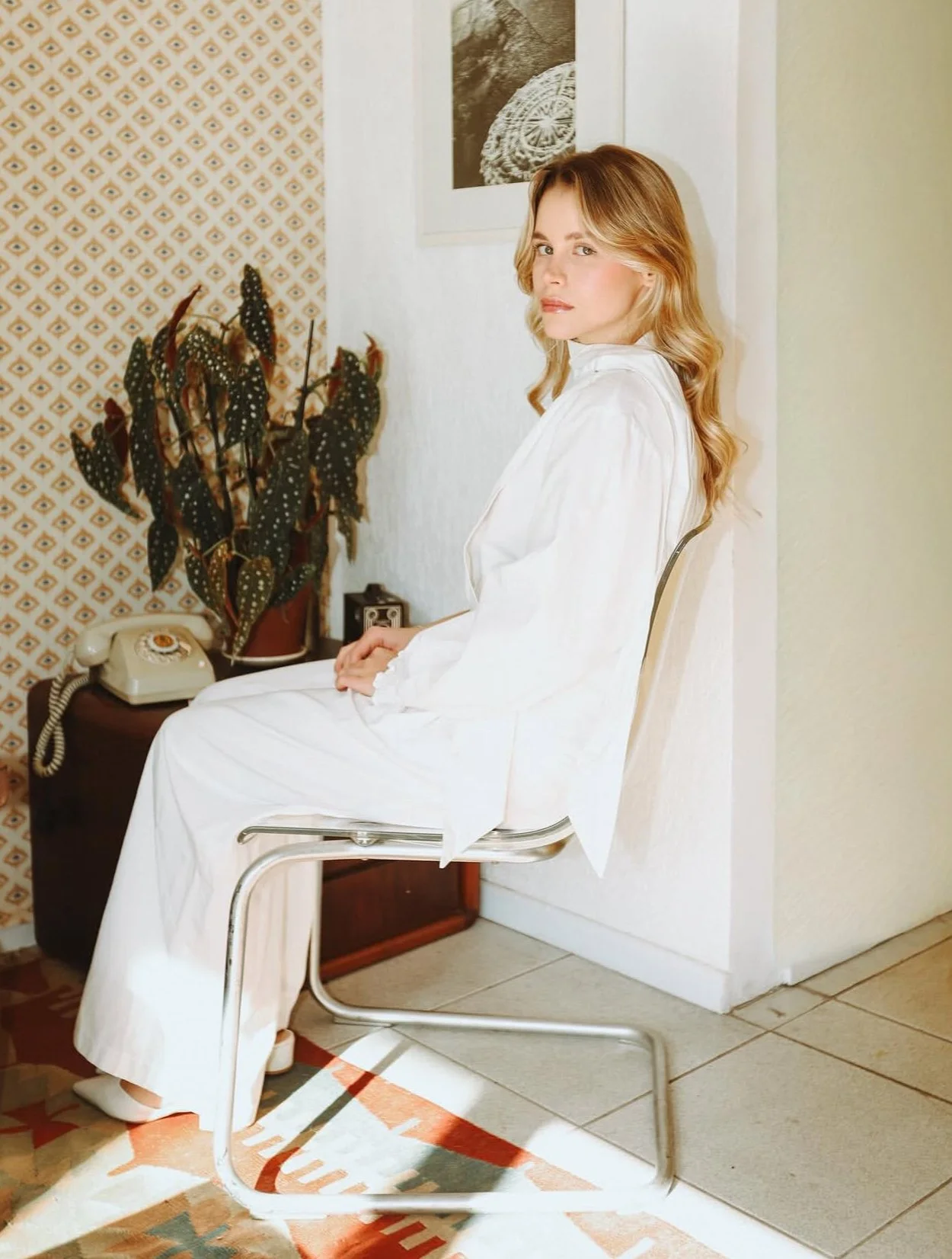 Mulher de cabelos loiros, vestindo roupa branca, sentada em uma cadeira na parede branca, com uma planta de folhas verdes ao fundo e um telefone antigo sobre uma mesa ao lado dela.