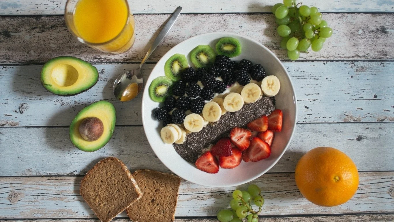 Breakfast plate with sliced bananas, strawberries, blackberries, and kiwi on a white dish. Surrounding the plate are two slices of bread, halves of an avocado, a glass of orange juice, a spoon, a bunch of green grapes, and an orange on a distressed wooden surface.