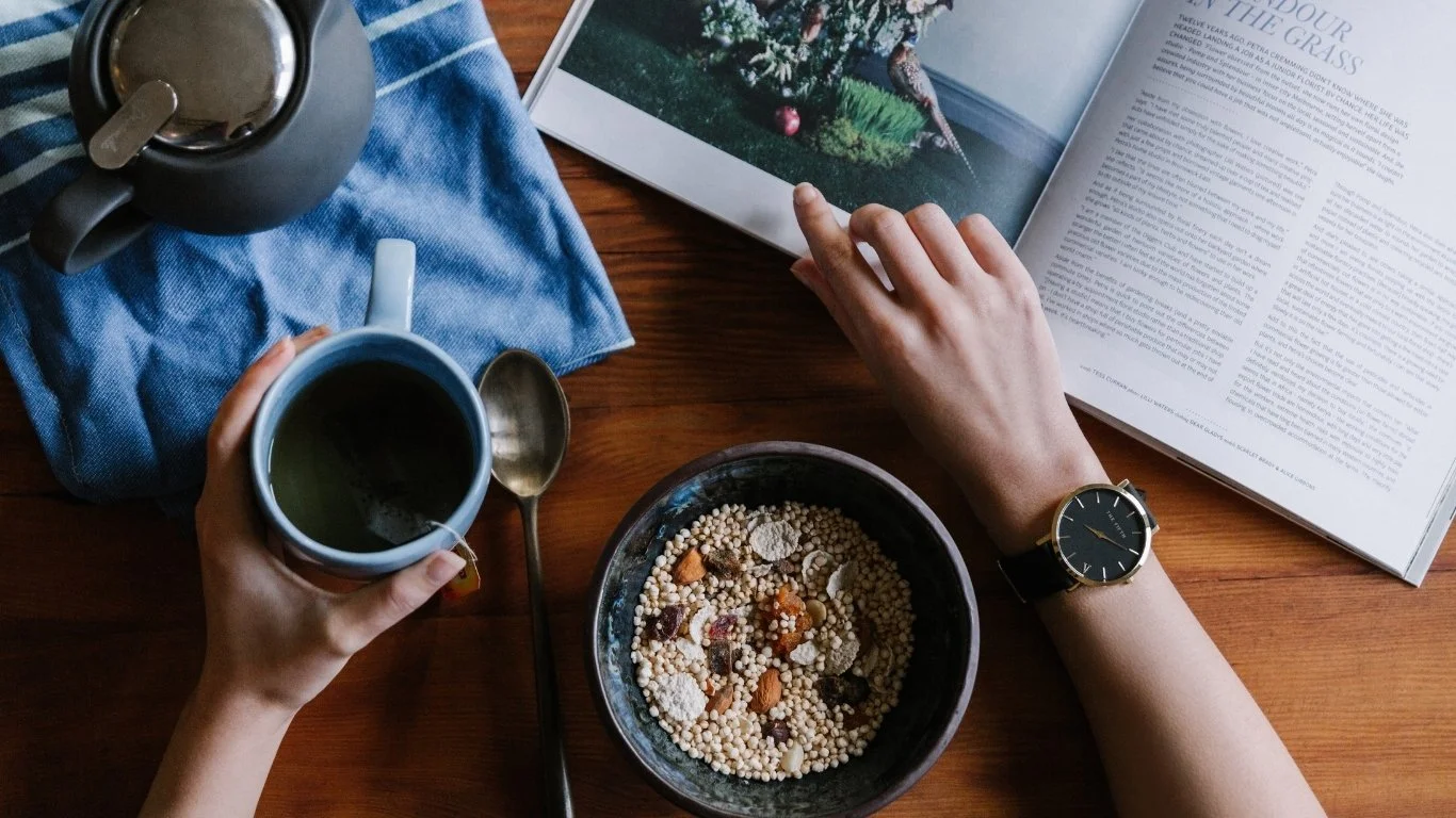 A person with a wristwatch reading a magazine at a wooden table. On the table are a bowl of cereal, a cup of tea or coffee, a spoon, a teapot, and an open magazine with a tree and Christmas decorations.