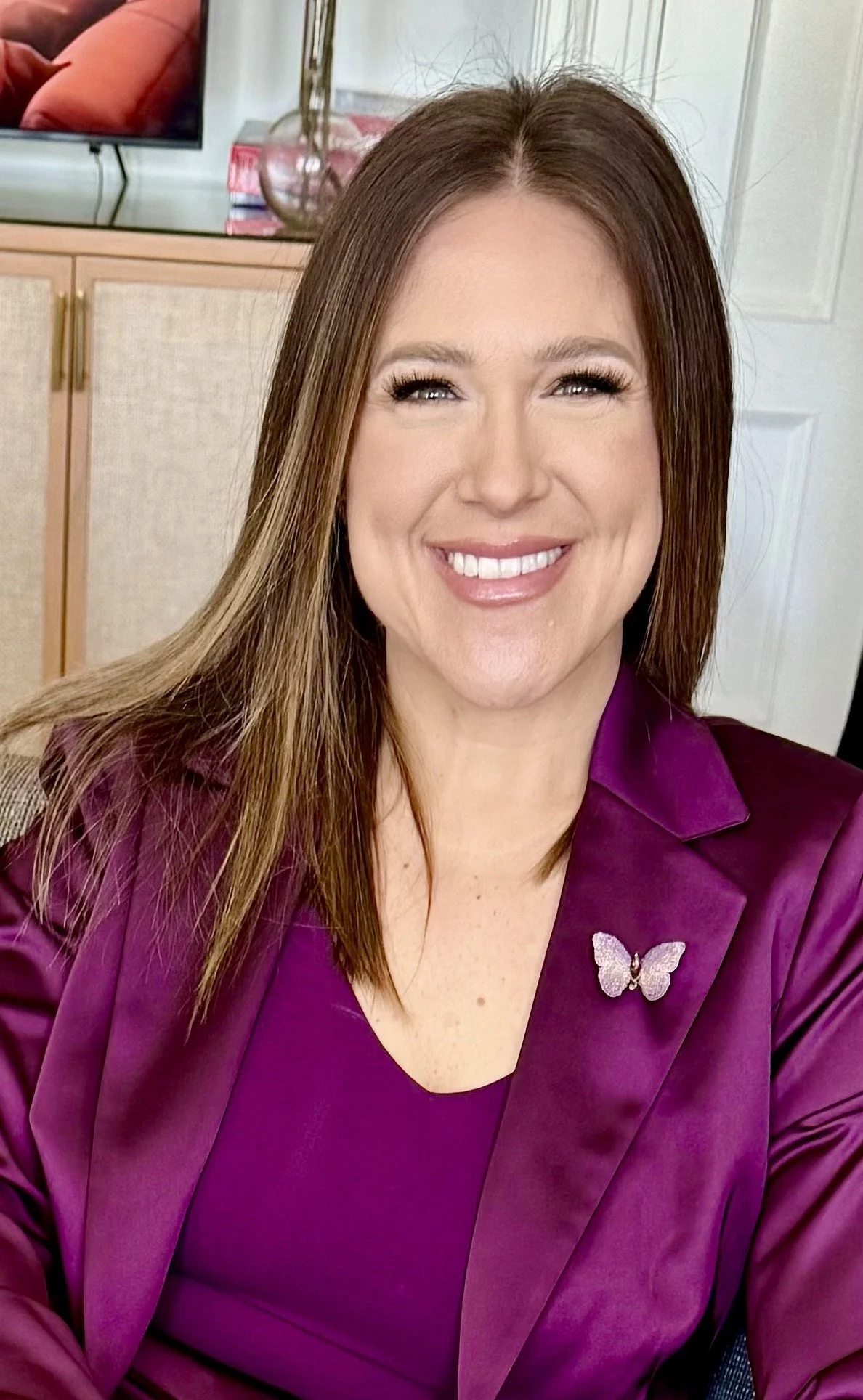 A woman with shoulder-length brown hair, wearing a purple blazer with a butterfly pin, smiling at the camera.