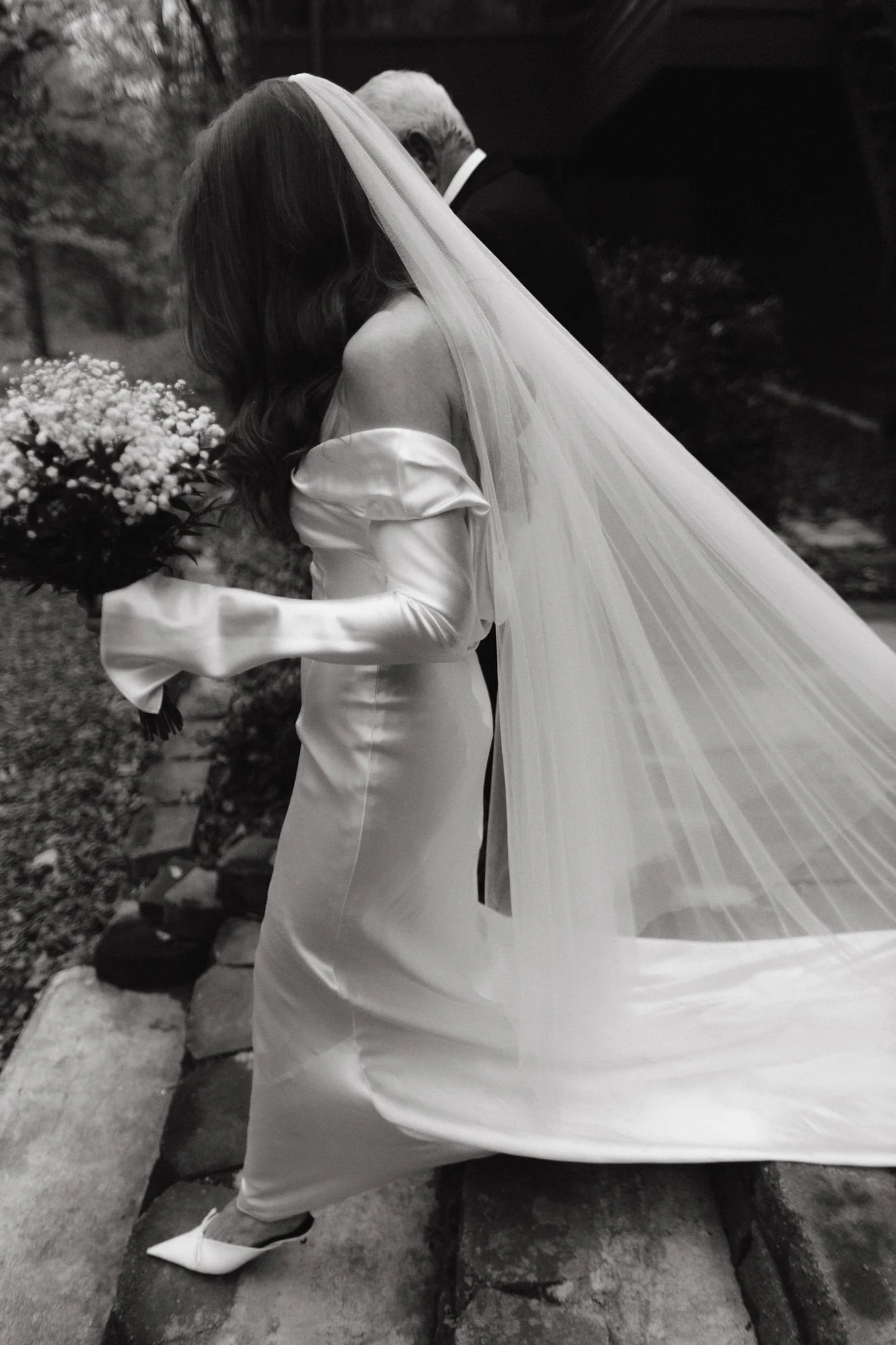 Bride in a silk off-the-shoulder gown and long flowing veil walking arm in arm with her father, photographed by Jenny Fu.