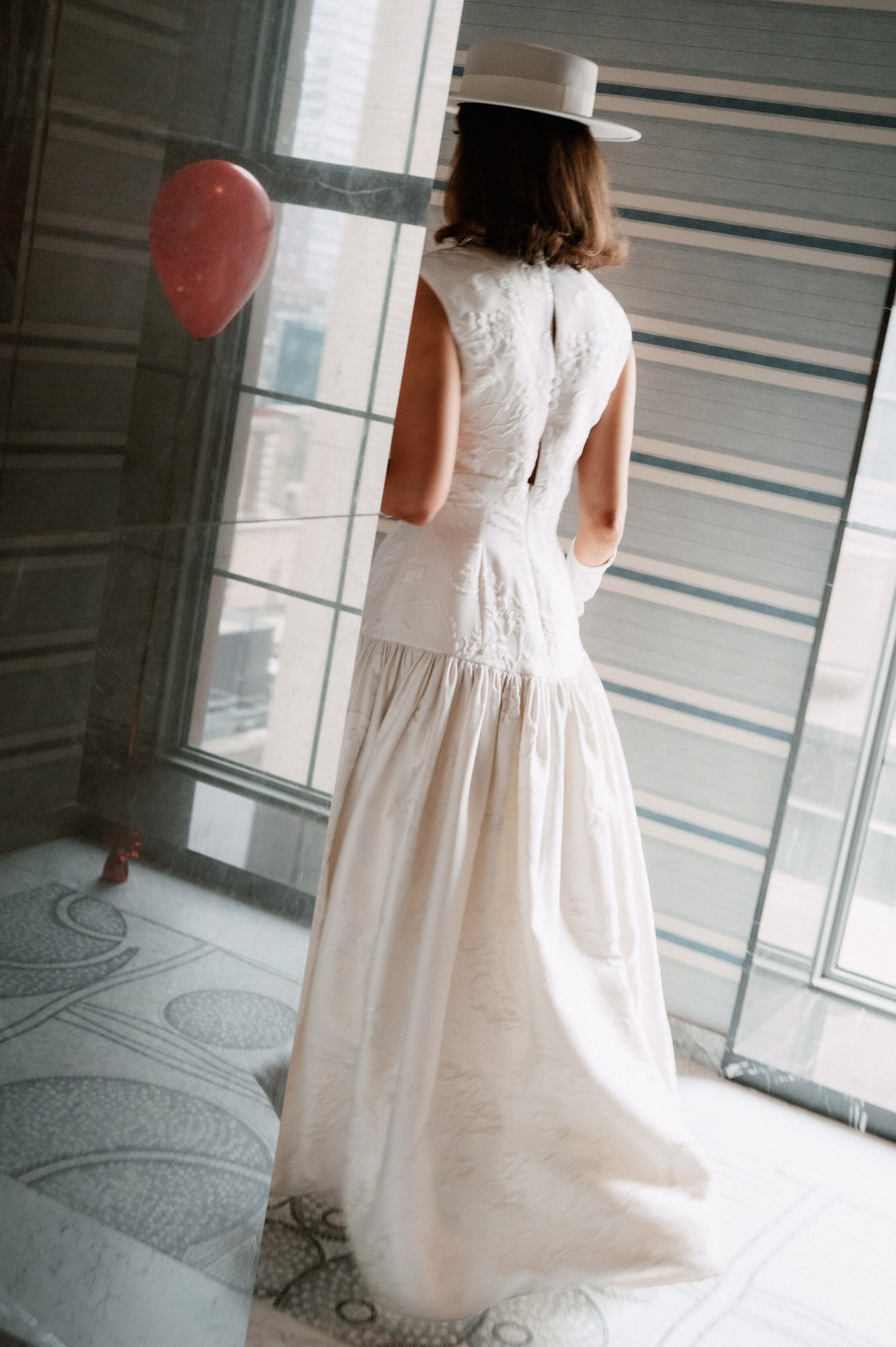 Bride in a boater hat and drop-waist gown seen from behind at a tall window with a red balloon drifting nearby at the Waldorf Astoria New York, photographed by Jenny Fu.