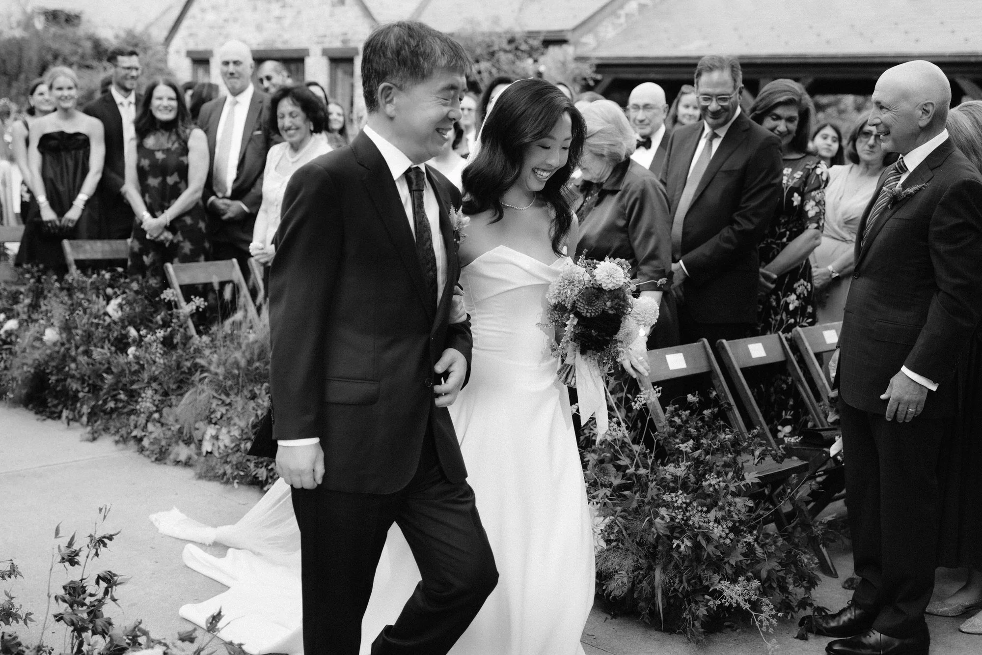 Bride walks down the aisle with her father through meadow-style florals in black and white at Blue Hill at Stone Barns, photographed by Jenny Fu.