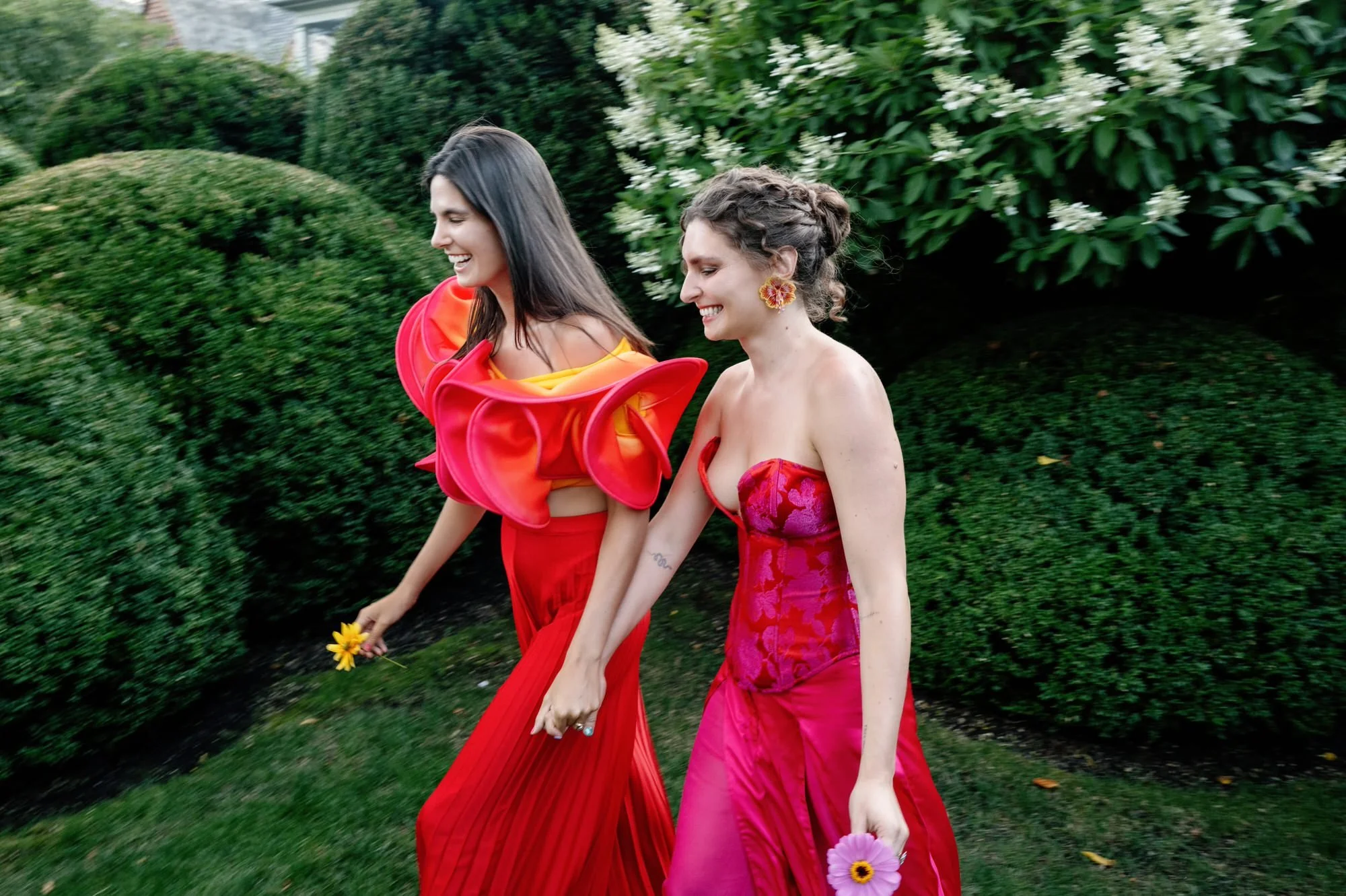 Two brides walking through clipped hedges, each carrying a single flower, at Valley Rock Inn in Sloatsburg, photographed by Jenny Fu.