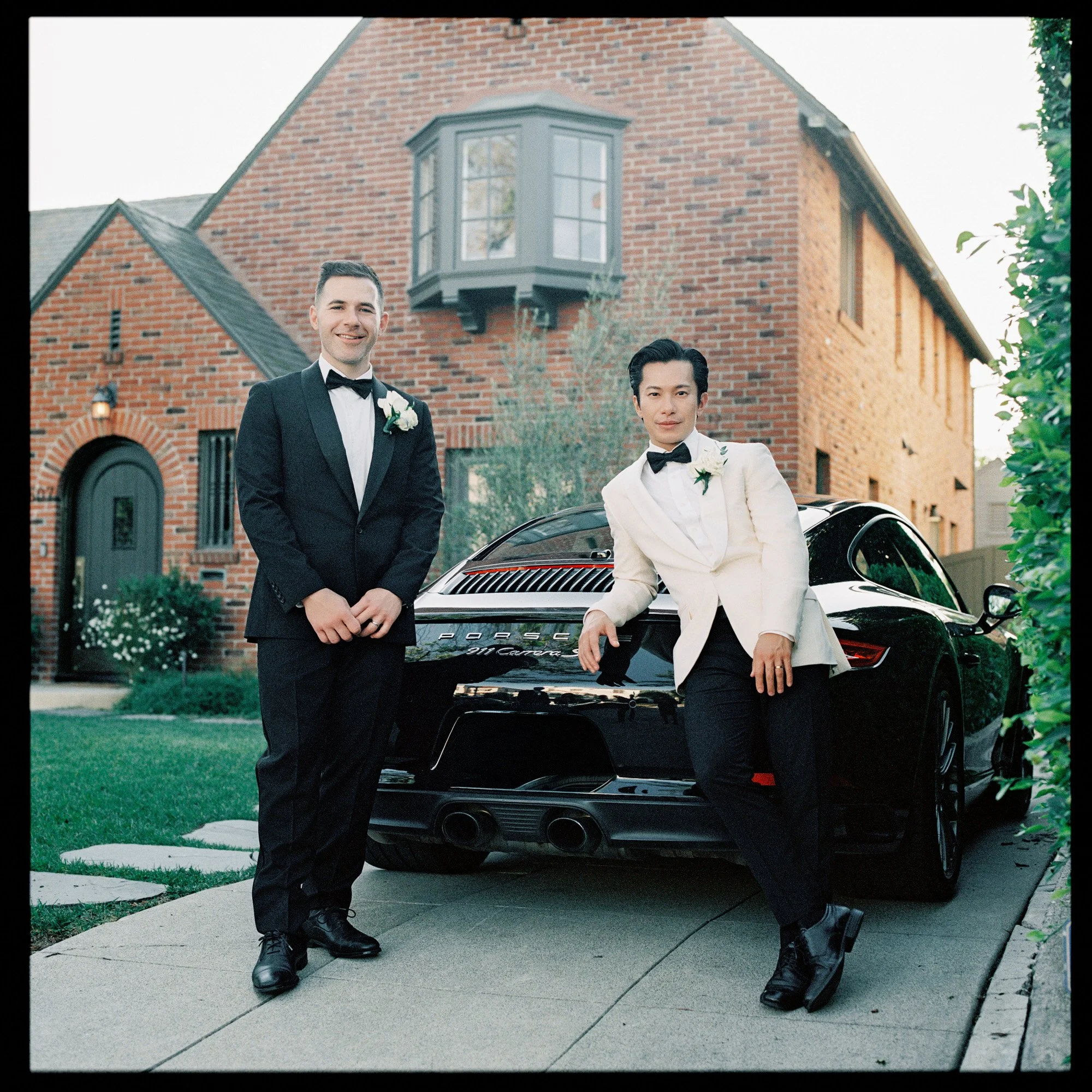 Two grooms in tuxedos beside a black Porsche outside a brick Tudor home in Los Angeles, photographed on film by Jenny Fu.