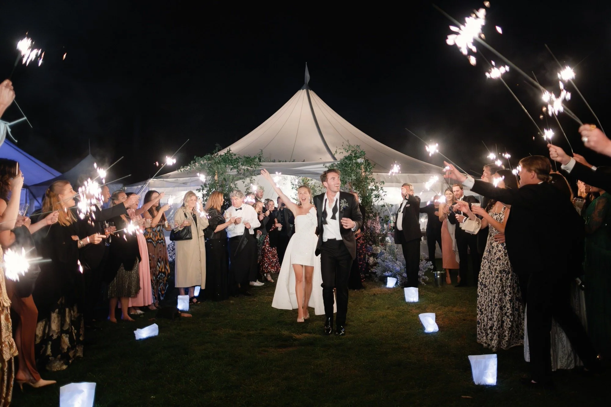 Bride and groom walking through a tunnel of sparklers held by guests outside the wedding tent