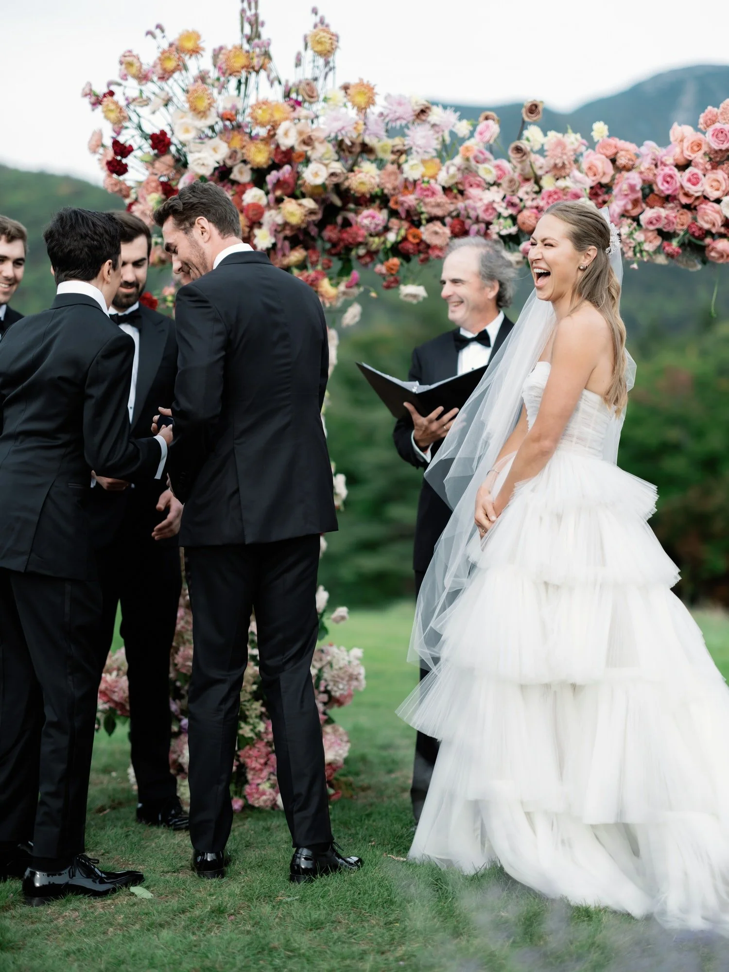 Bride laughing joyfully during wedding ceremony under floral arch at The Ausable Club in the Adirondacks