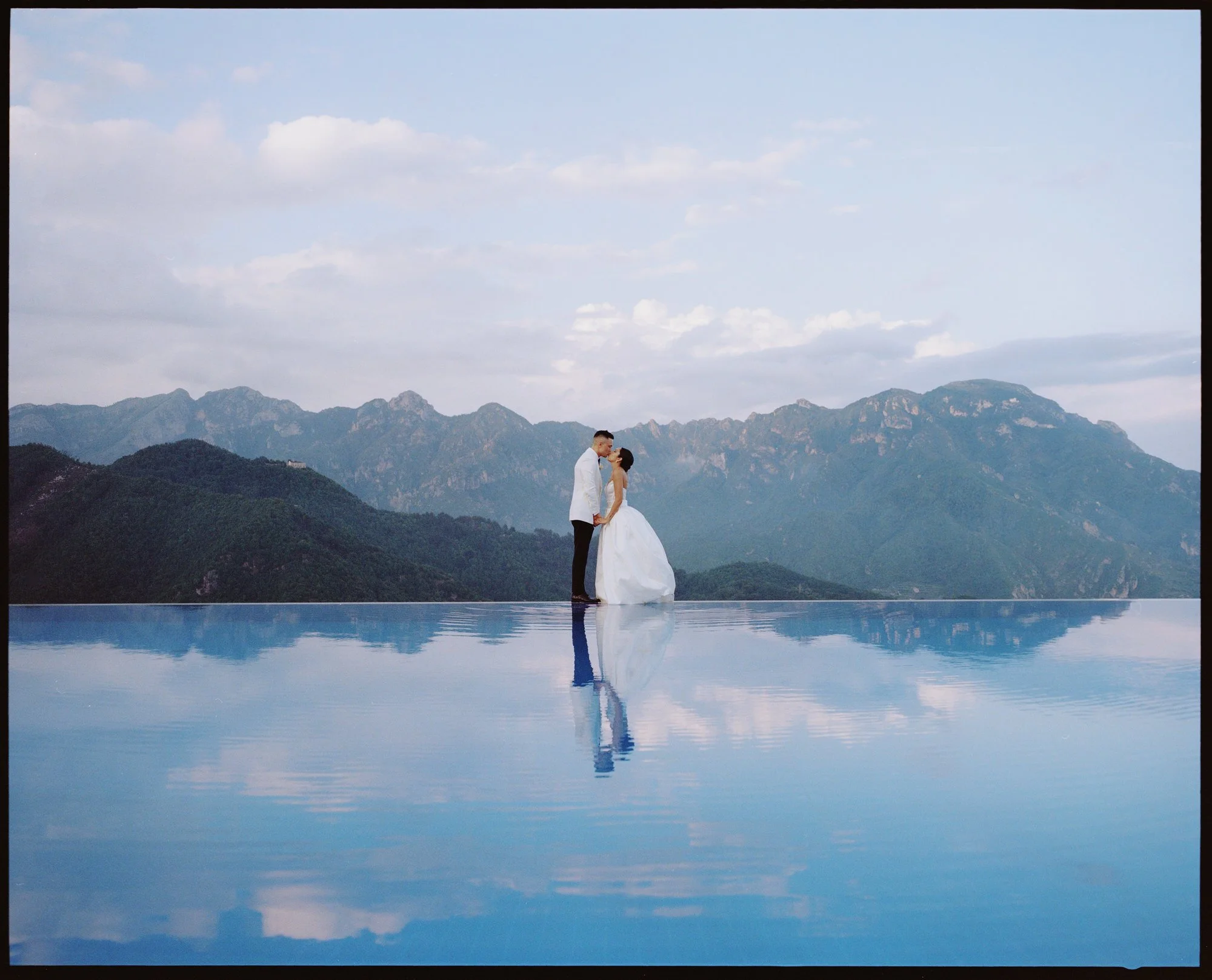 Bride and groom kissing by the iconic infinity pool reflecting the mountains at Caruso, A Belmond Hotel in Ravello, Italy, photographed by Jenny Fu.