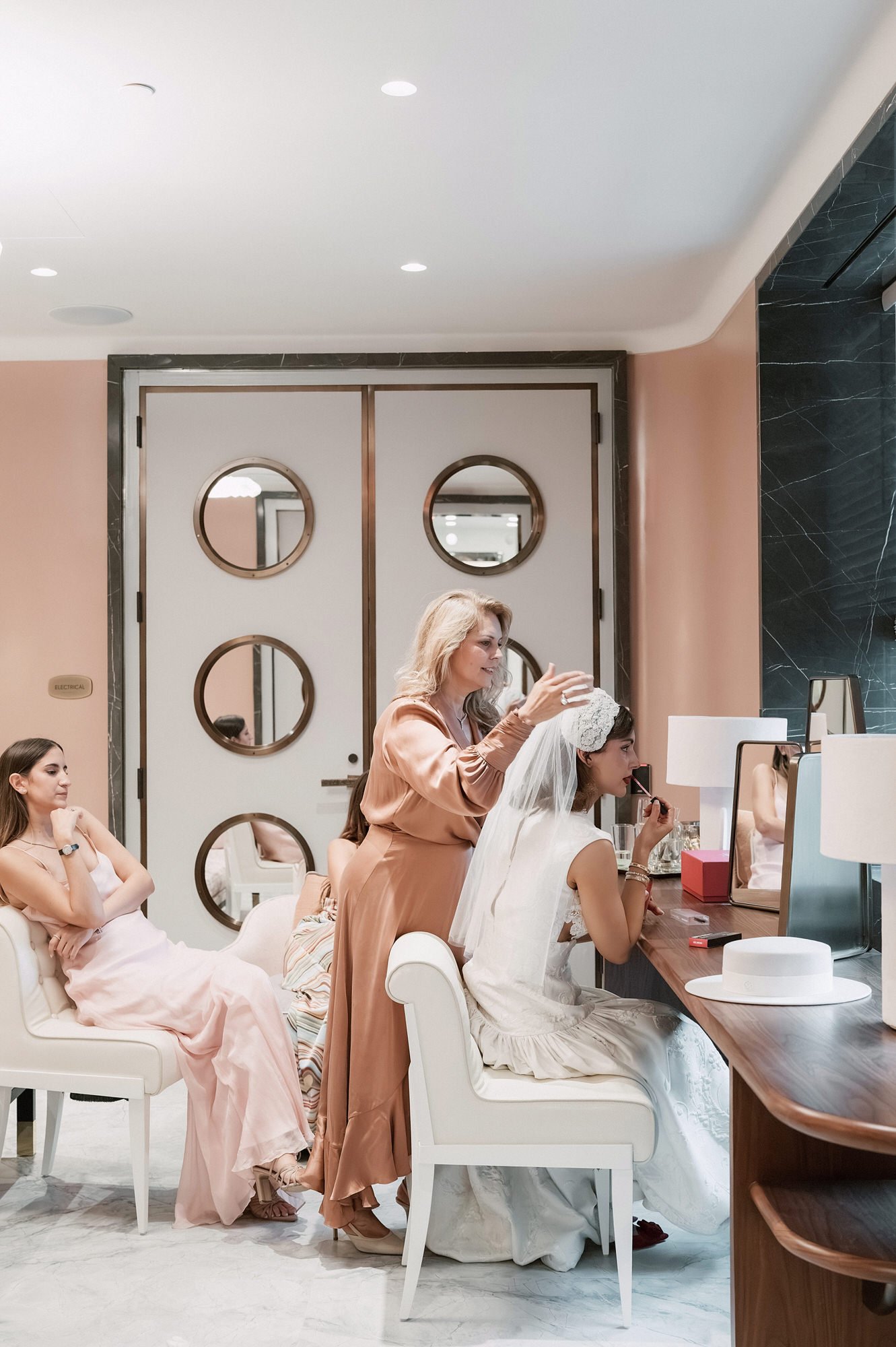Bride having her veil adjusted at a vanity while bridesmaids look on in a blush suite at the Waldorf Astoria New York, photographed by Jenny Fu.