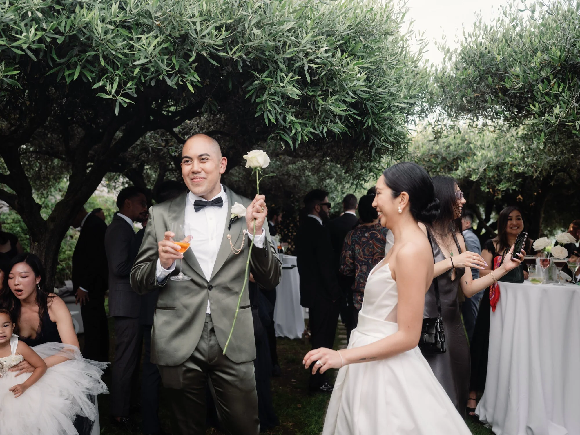 Bride and officiant sharing a joyful moment with drinks in hand under olive trees at Caruso, A Belmond Hotel in Ravello, Italy, photographed by Jenny Fu.