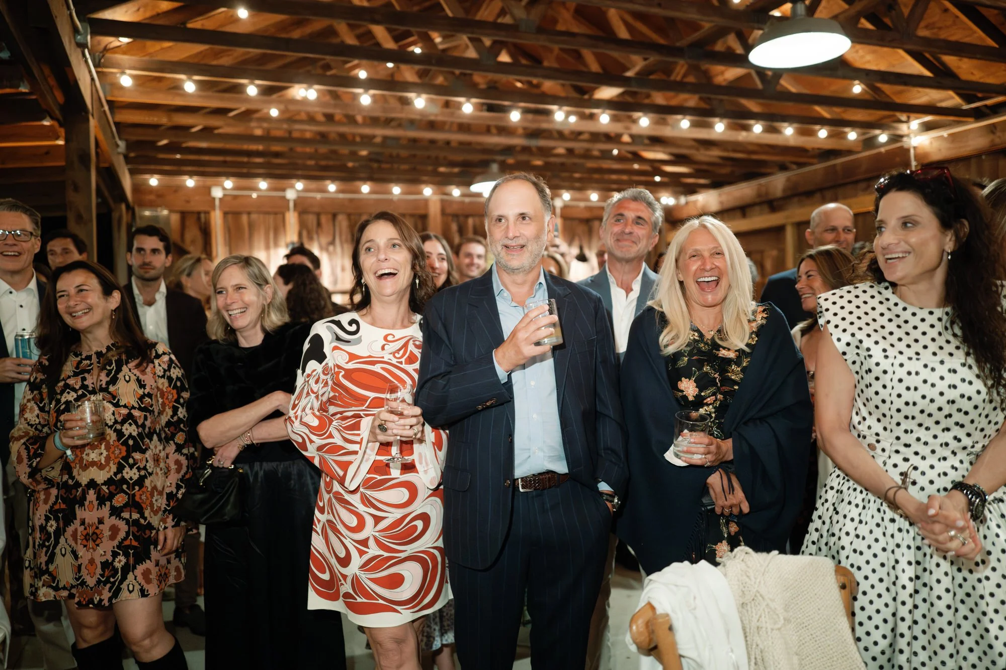 Guests laughing and reacting together under barn string lights during the rehearsal dinner at Troutbeck in Amenia, New York, photographed by Jenny Fu.