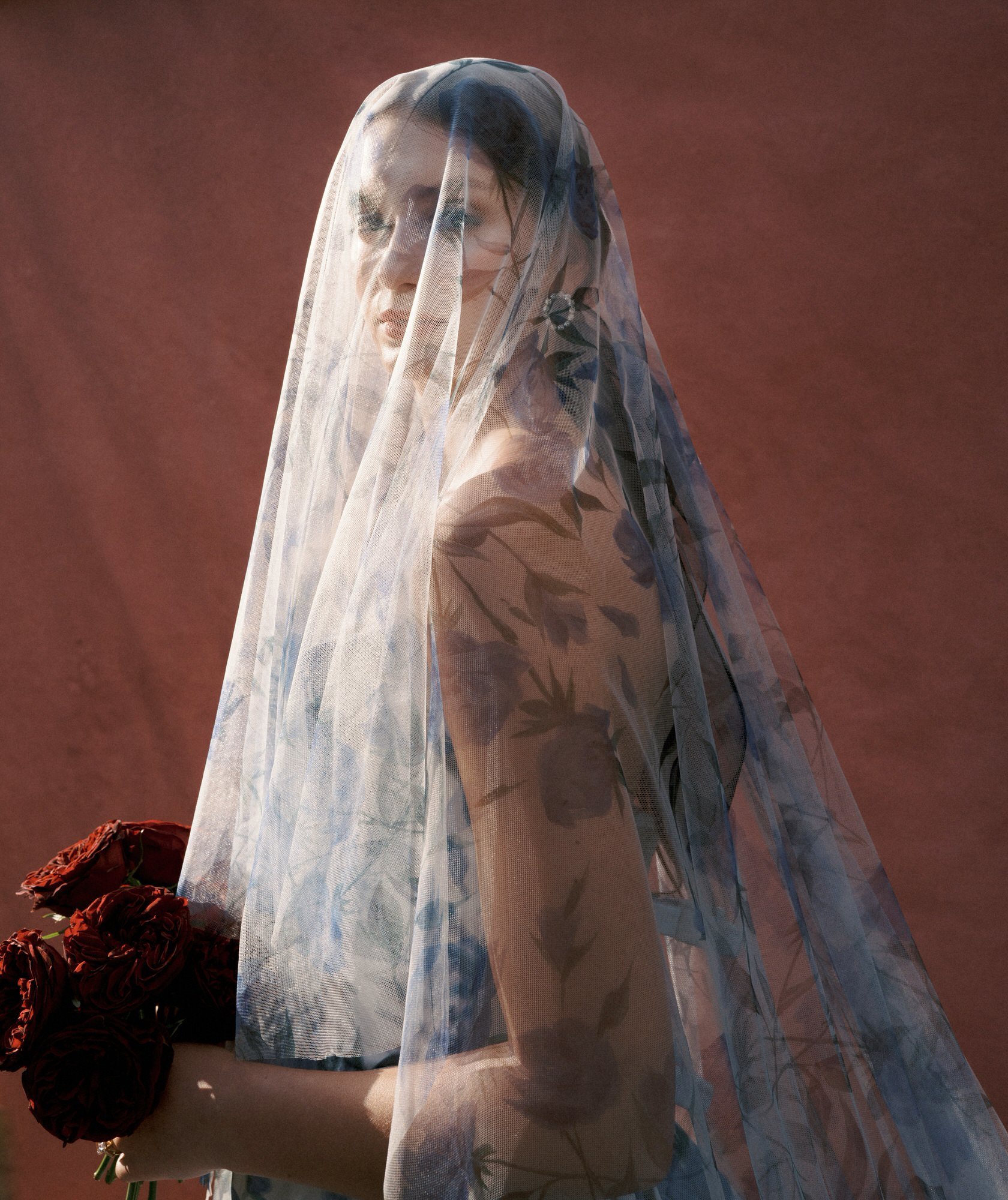 Bride draped in a floral-patterned veil holding red roses against a terracotta wall, photographed by Jenny Fu.