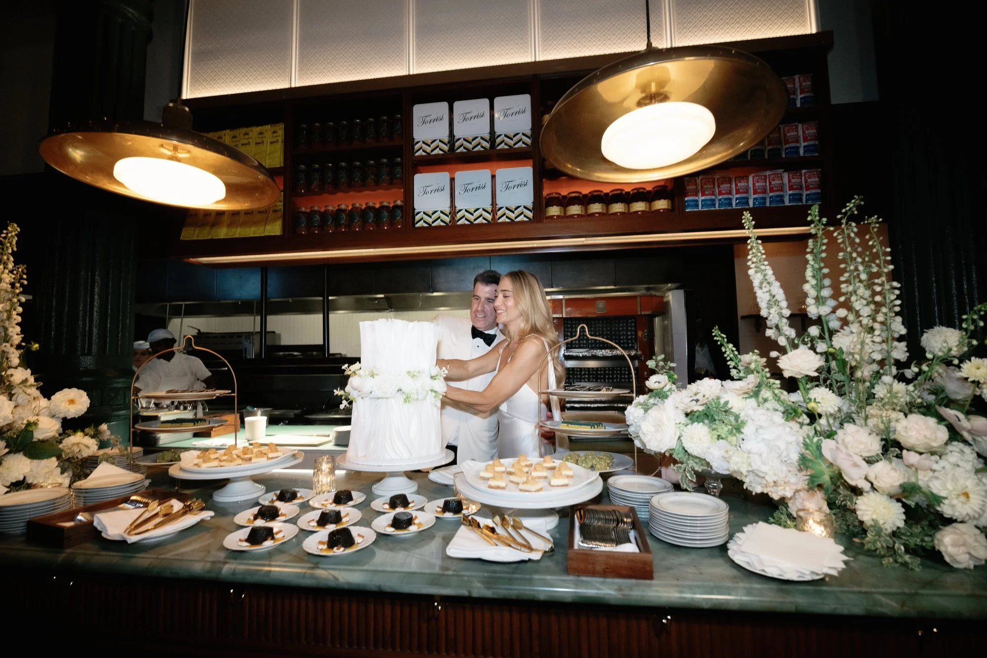 Bride and groom cutting their elegant white tiered cake surrounded by desserts and flowers at Torrisi in Manhattan, photographed by Jenny Fu.