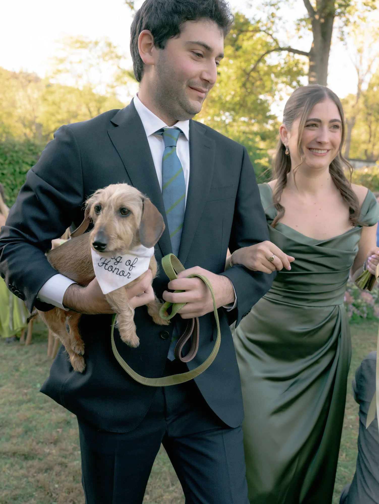 Small dachshund wearing a "dog of honor" bandana held by a groomsman with bridesmaid smiling beside him at Troutbeck in Amenia, New York, photographed by Jenny Fu.