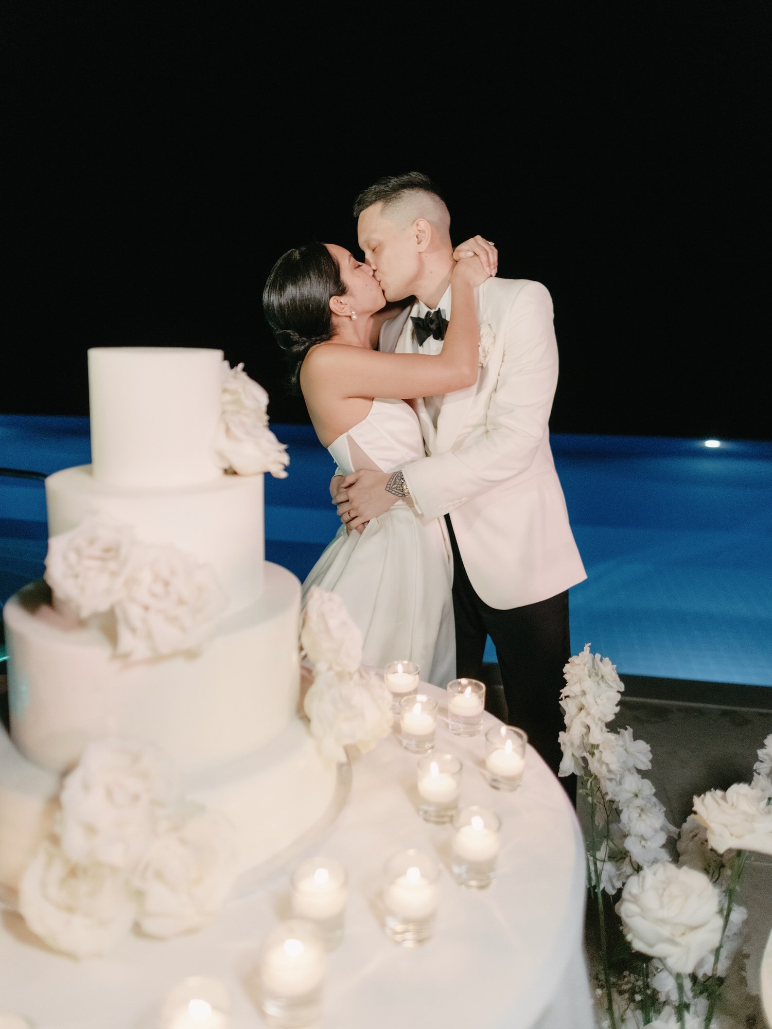 Bride and groom sharing a kiss beside their candlelit white wedding cake by the pool at Caruso, A Belmond Hotel in Ravello, Italy, photographed by Jenny Fu.