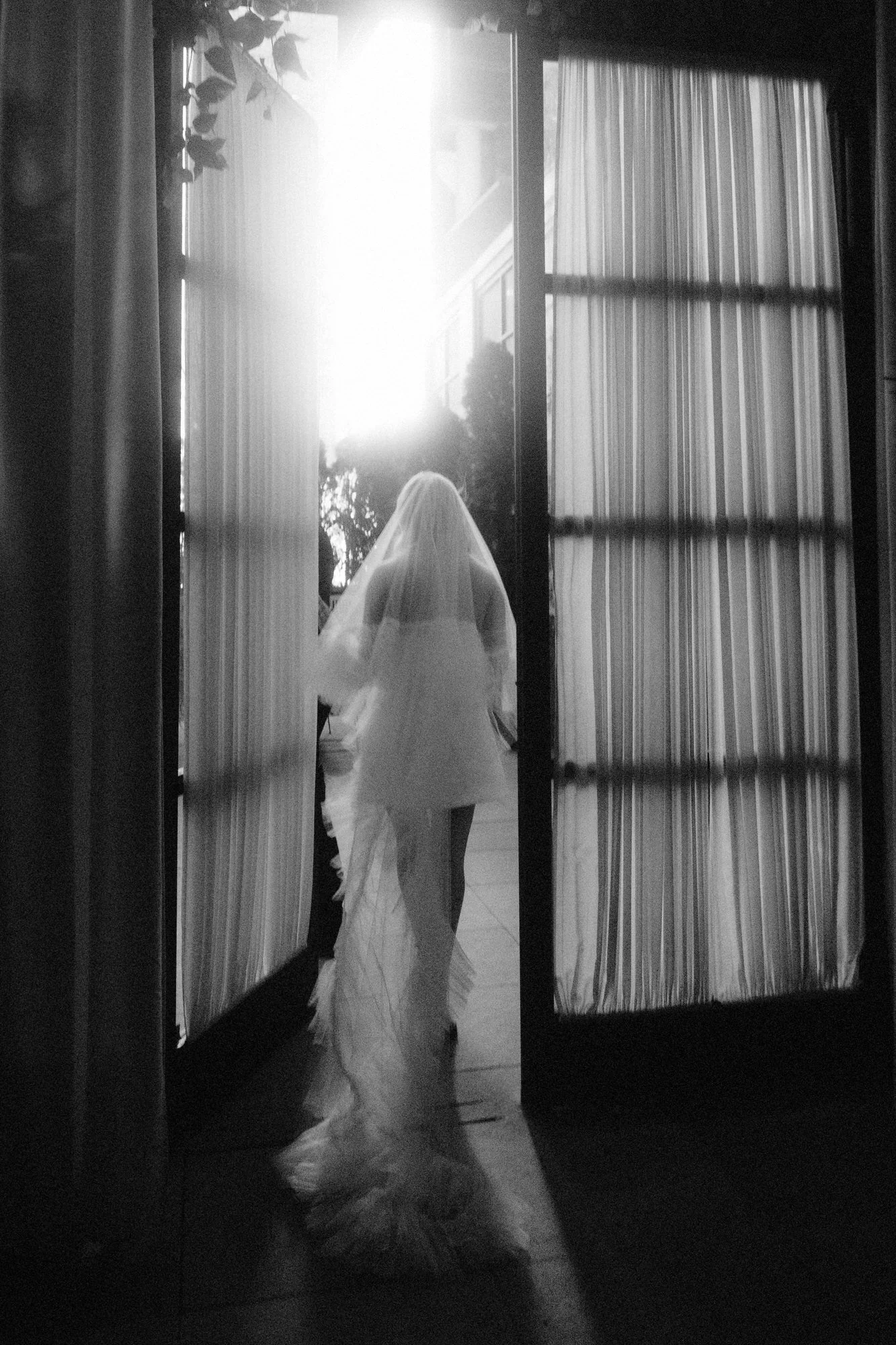 Bride walking through open French doors into glowing sunset light, her veil trailing behind her, photographed by Jenny Fu.