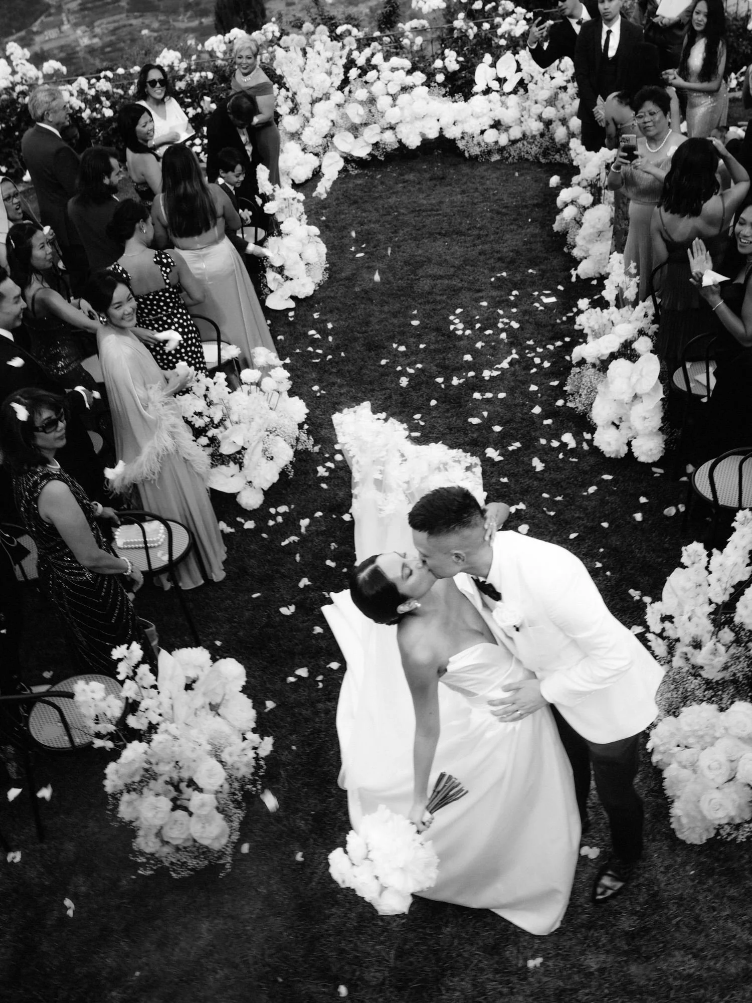 Black-and-white image of the bride and groom sharing a kiss down the aisle surrounded by cheering guests at Caruso, A Belmond Hotel in Ravello, Italy, photographed by Jenny Fu.