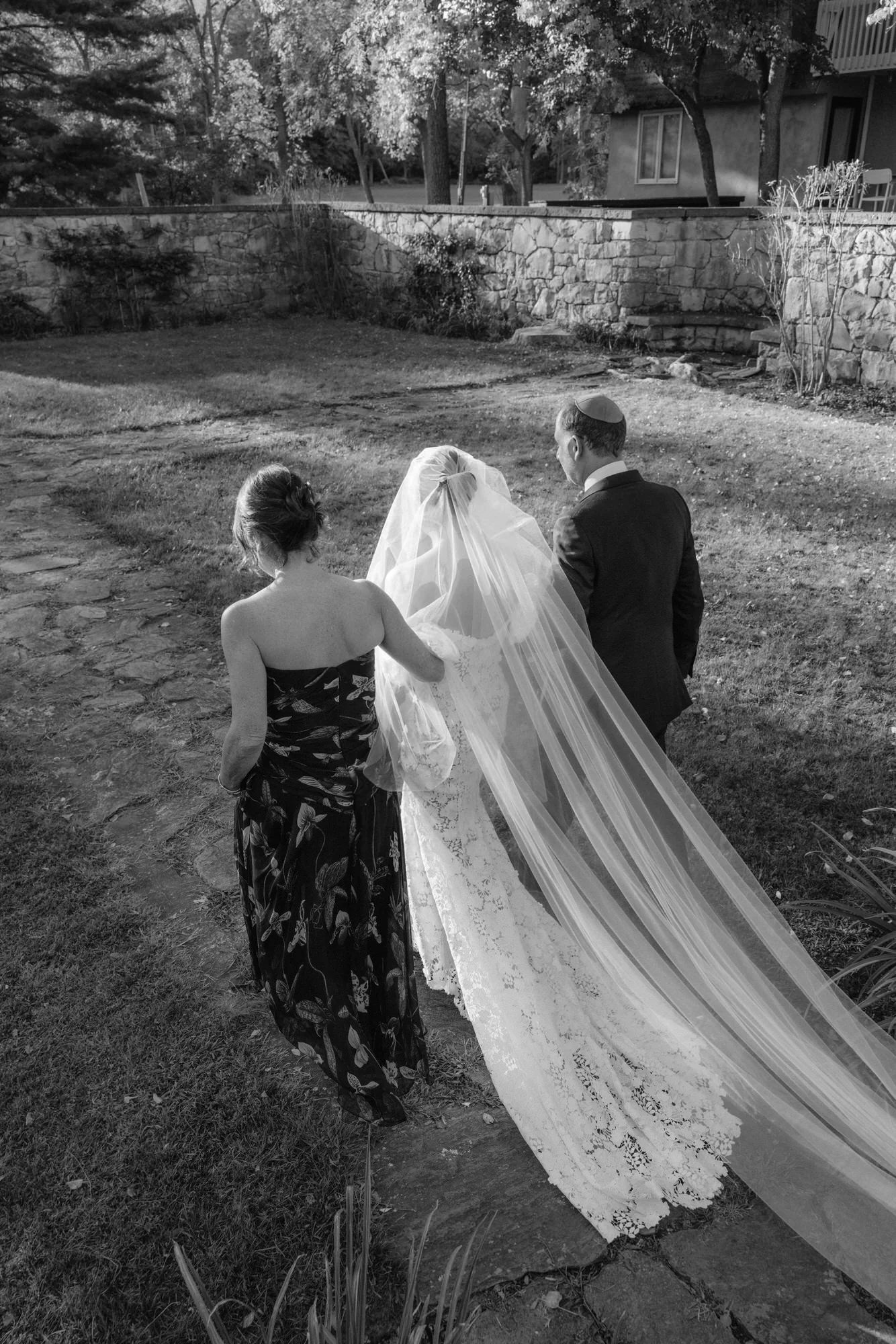 Bride walking arm-in-arm with her parents along a stone path toward the ceremony at Troutbeck in Amenia, photographed by Jenny Fu.