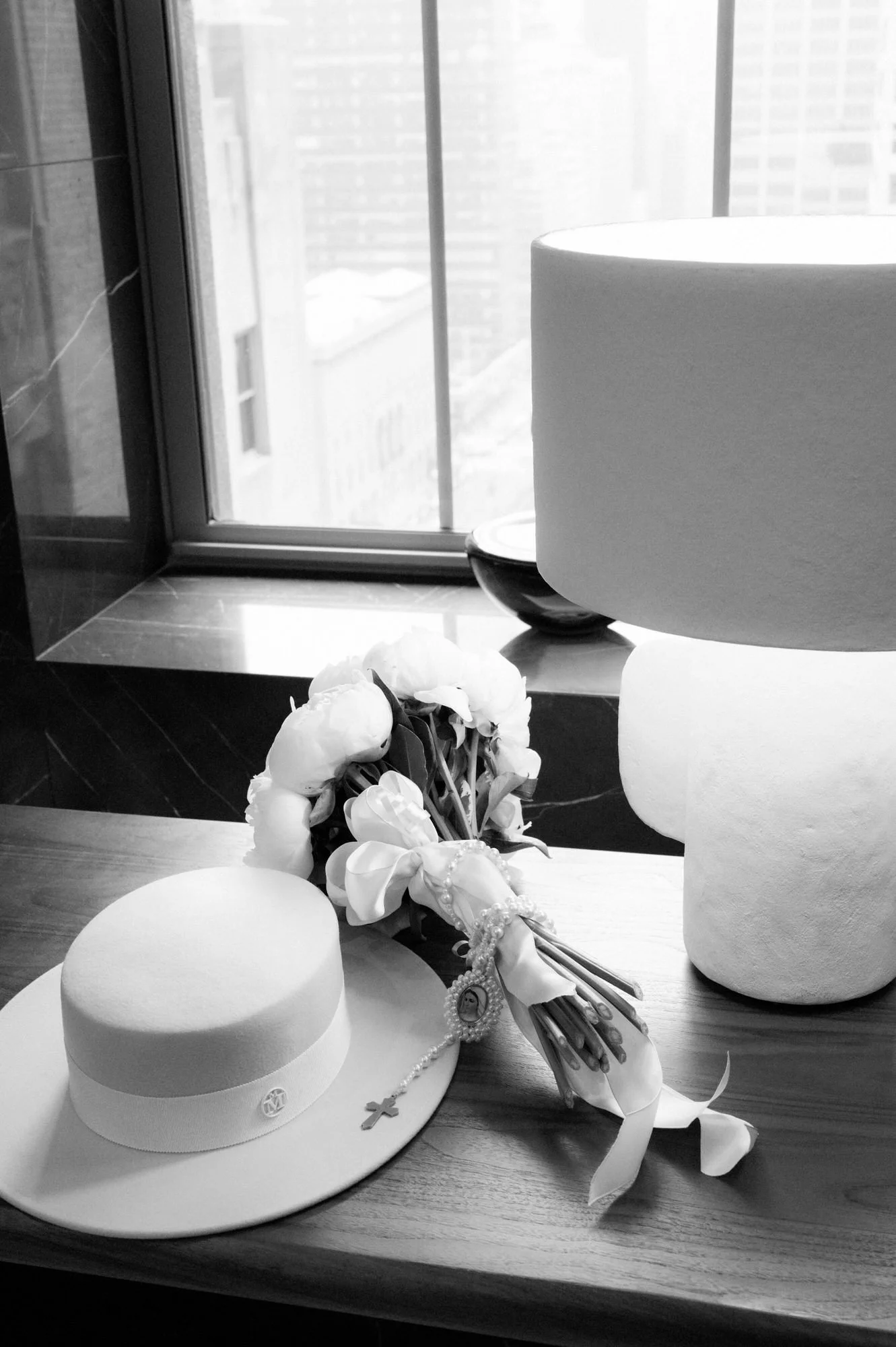 Black and white still life of the bride's boater hat and white bouquet beside a lamp at a window at the Waldorf Astoria New York, photographed by Jenny Fu.