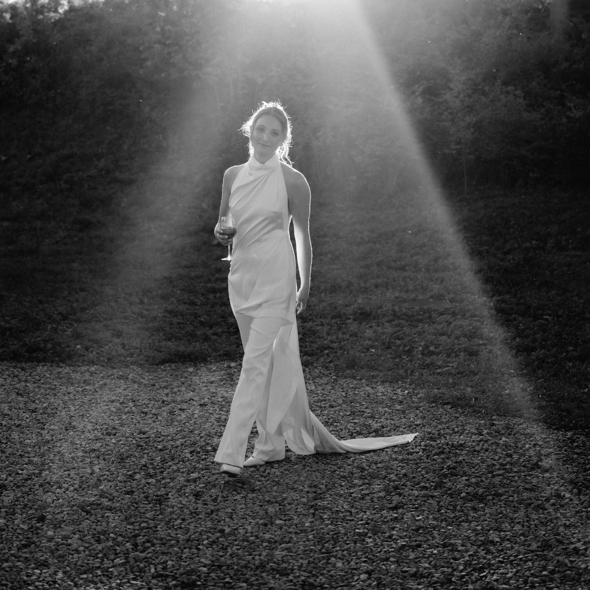 Bride walking across gravel with a glass of wine as sunlight beams through the trees at Troutbeck in Amenia, photographed by Jenny Fu.