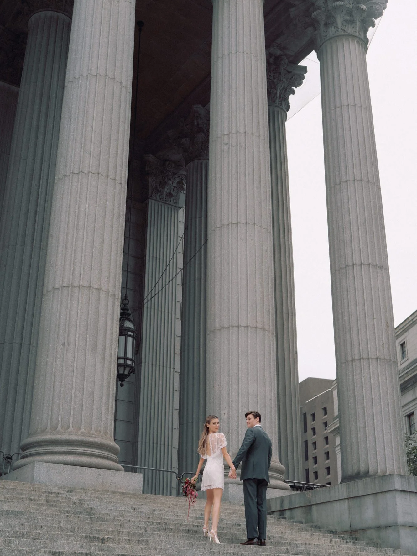 Bride and groom standing small beneath towering stone columns on the courthouse steps in Lower Manhattan, photographed by Jenny Fu.