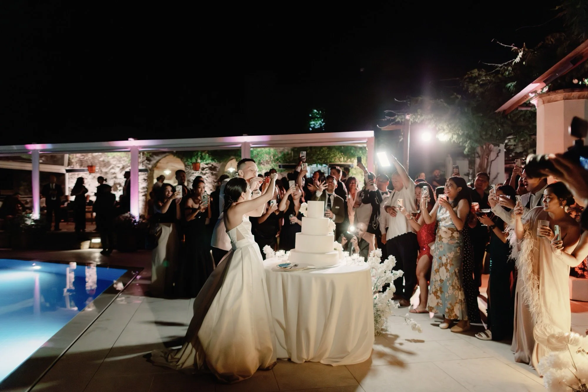 Bride and groom cutting their wedding cake surrounded by cheering guests at Caruso, A Belmond Hotel in Ravello, Italy, photographed by Jenny Fu.