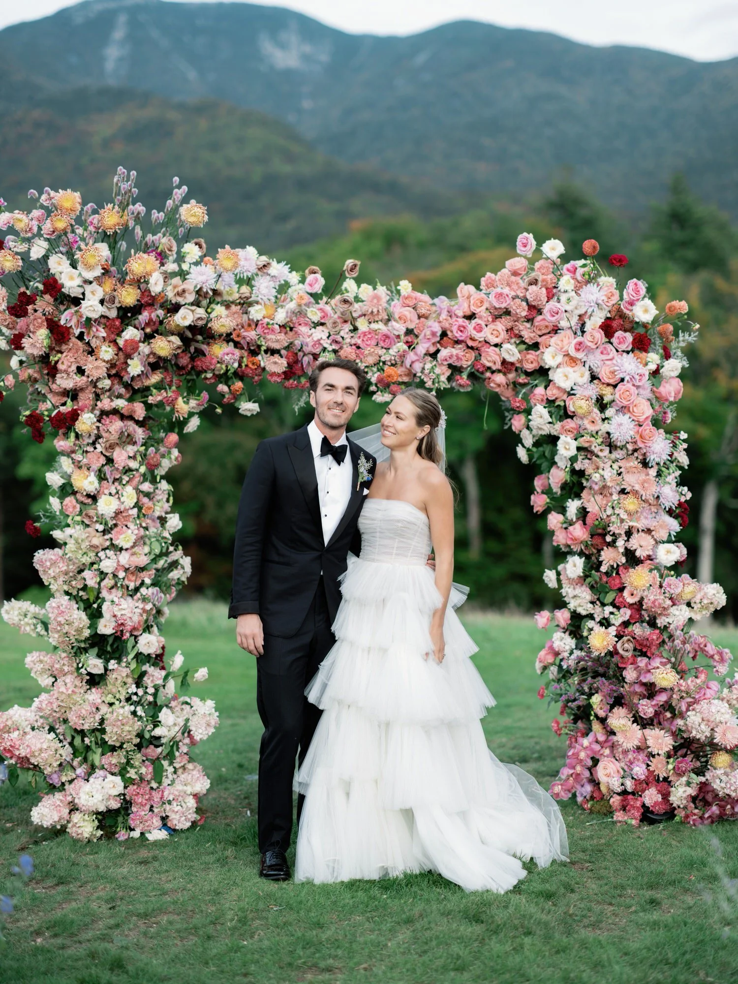Olivia and Jack smiling together under a colorful floral arch at their wedding ceremony at the Ausable Club in the Adirondacks