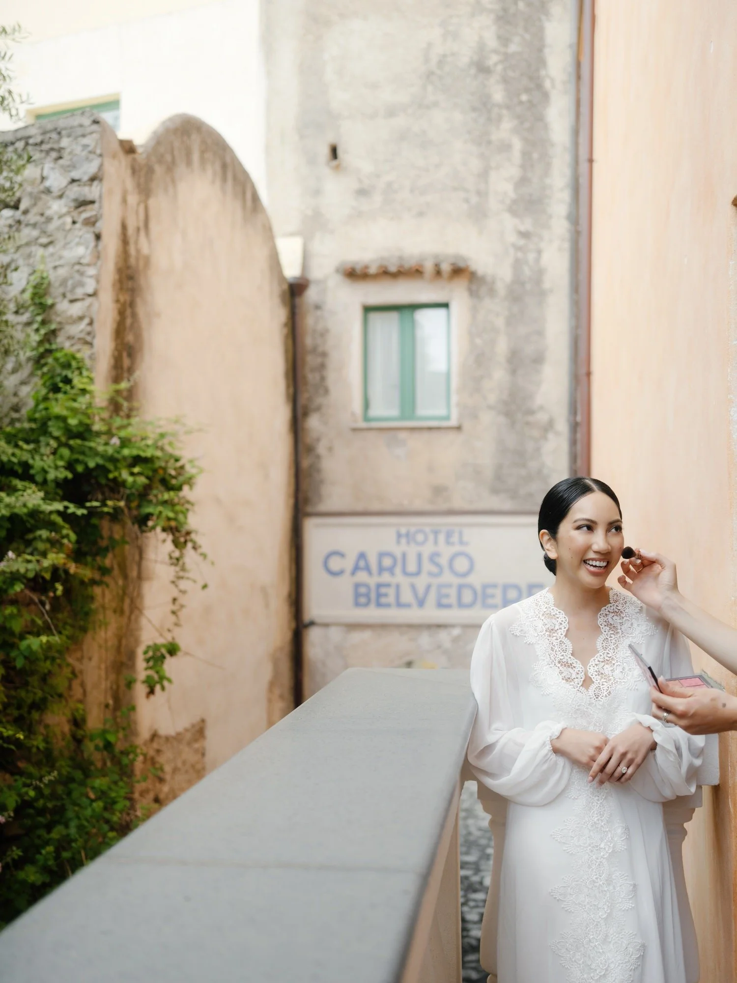 Bride smiling as a makeup artist adjusts her lipstick outside Hotel Caruso Belvedere in Ravello, Italy, photographed by Jenny Fu.