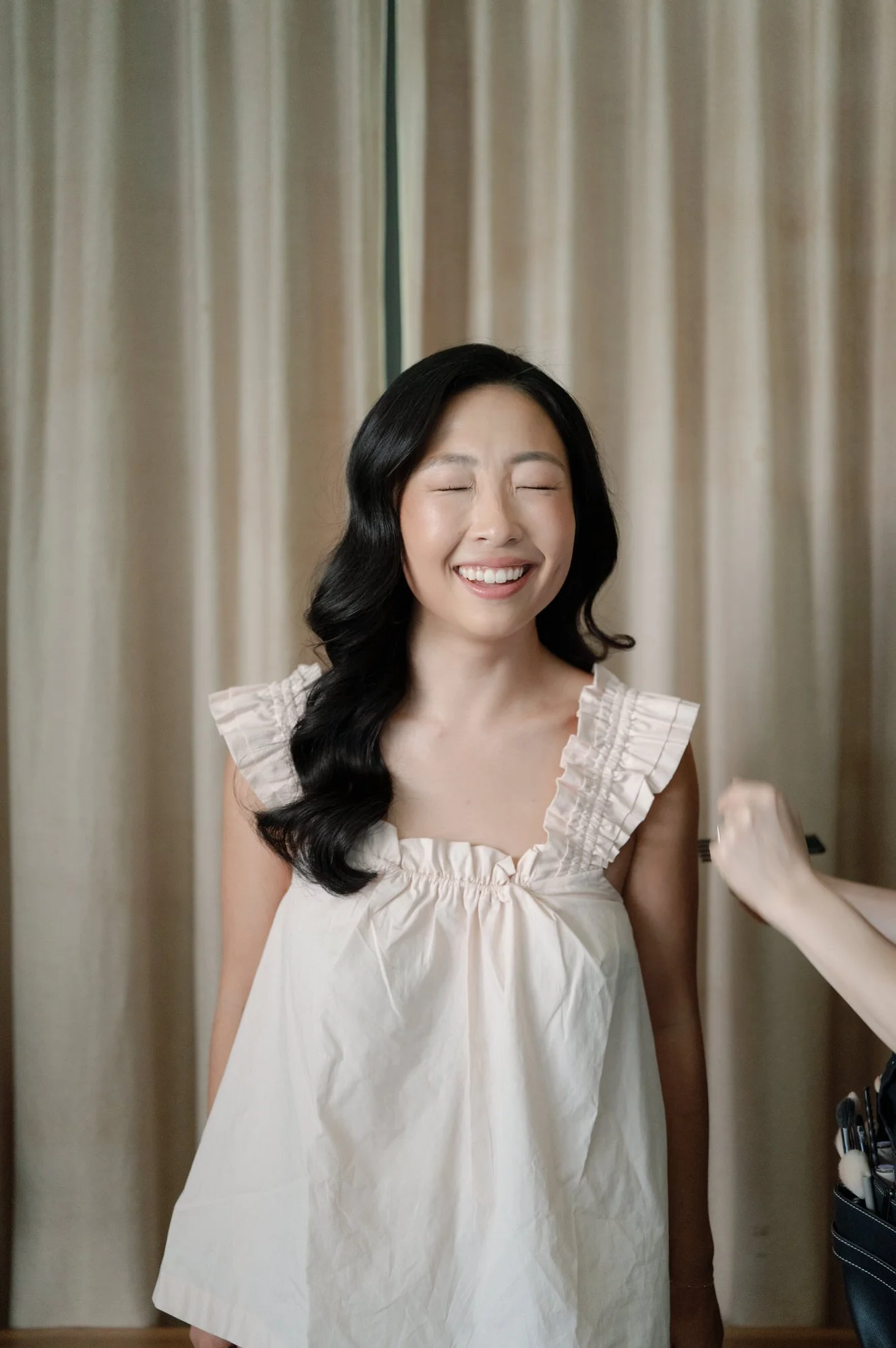 Bride smiling with eyes closed as a stylist finishes hair and makeup against soft drapery at Blue Hill at Stone Barns, photographed by Jenny Fu.