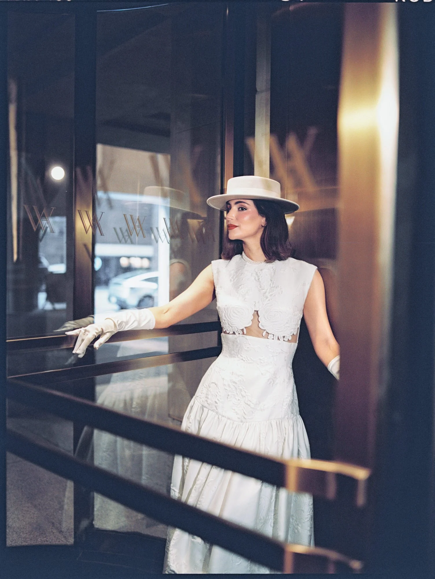 Bride in a white boater hat and two-piece gown leaning on a brass railing at the Waldorf Astoria New York, photographed by Jenny Fu.