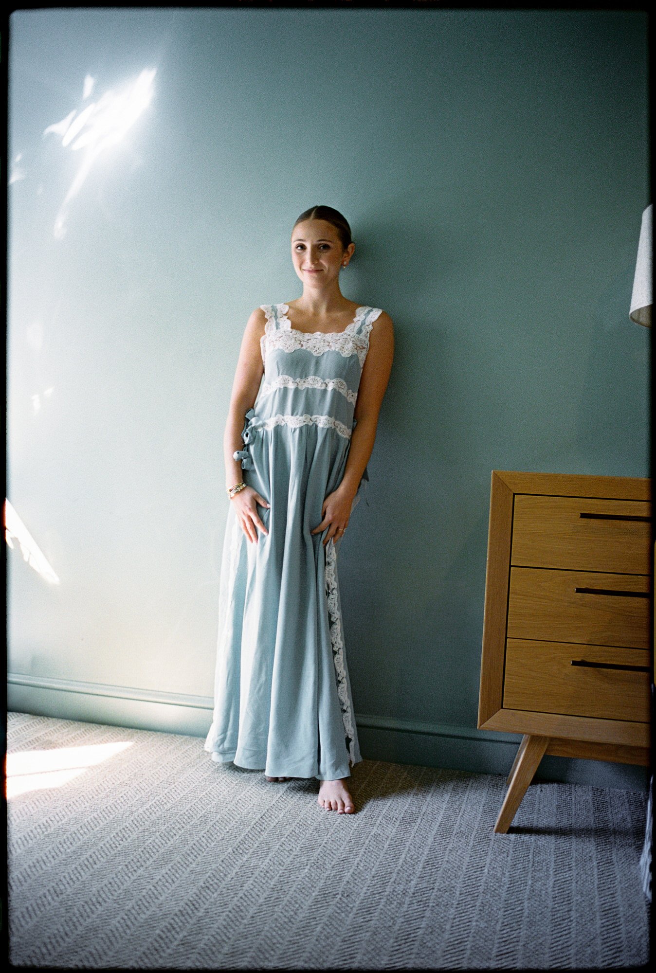 Bride posing barefoot in a soft blue lace-trimmed slip dress against a pale green wall at Troutbeck in Amenia, photographed by Jenny Fu.