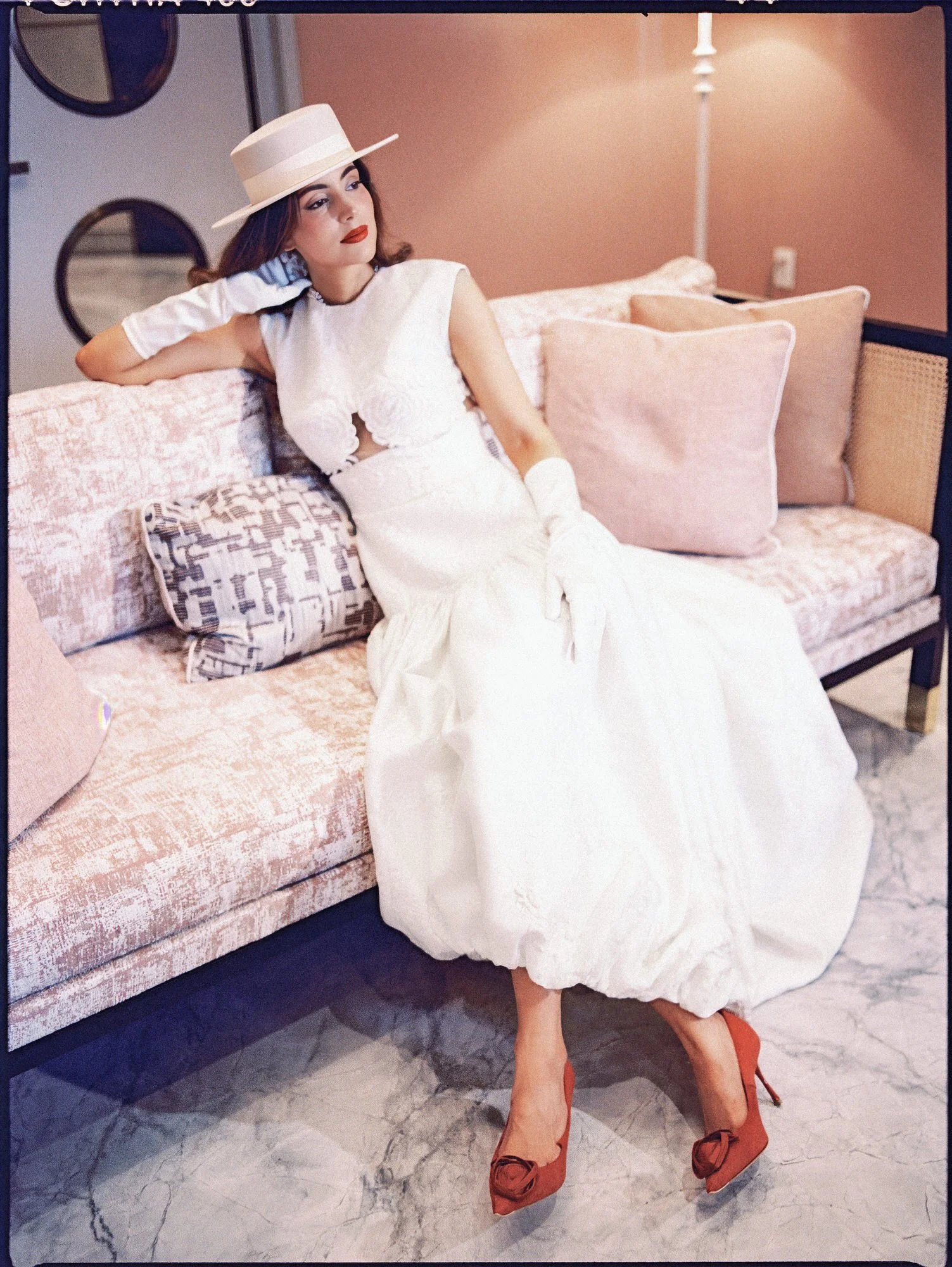 Bride in a white boater hat, gloves and red heels reclining on a sofa with one hand raised to her hat at the Waldorf Astoria New York, photographed by Jenny Fu.