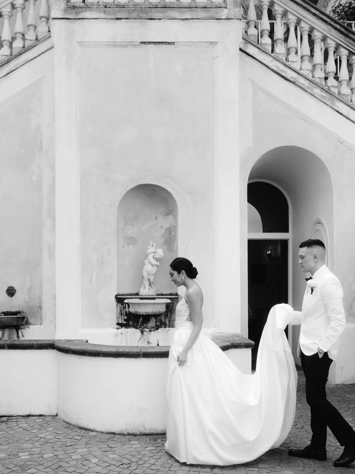 Groom gently holding the bride’s train as they walk past a historic fountain at Caruso, A Belmond Hotel in Ravello, Italy, photographed by Jenny Fu.
