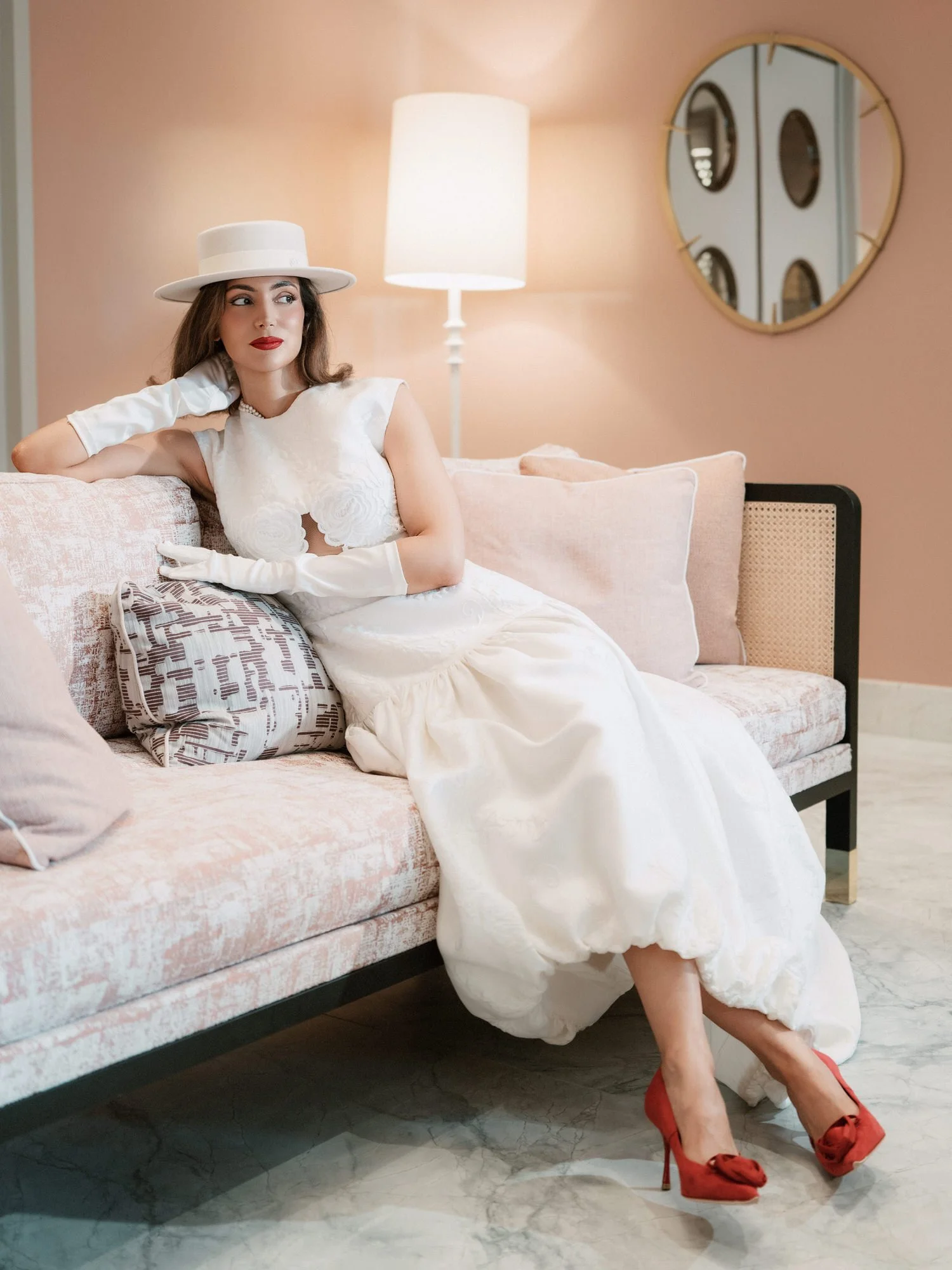 Bride in a white boater hat, gloves and red heels lounging on a blush sofa beneath a round mirror in a suite at the Waldorf Astoria New York, photographed by Jenny Fu.