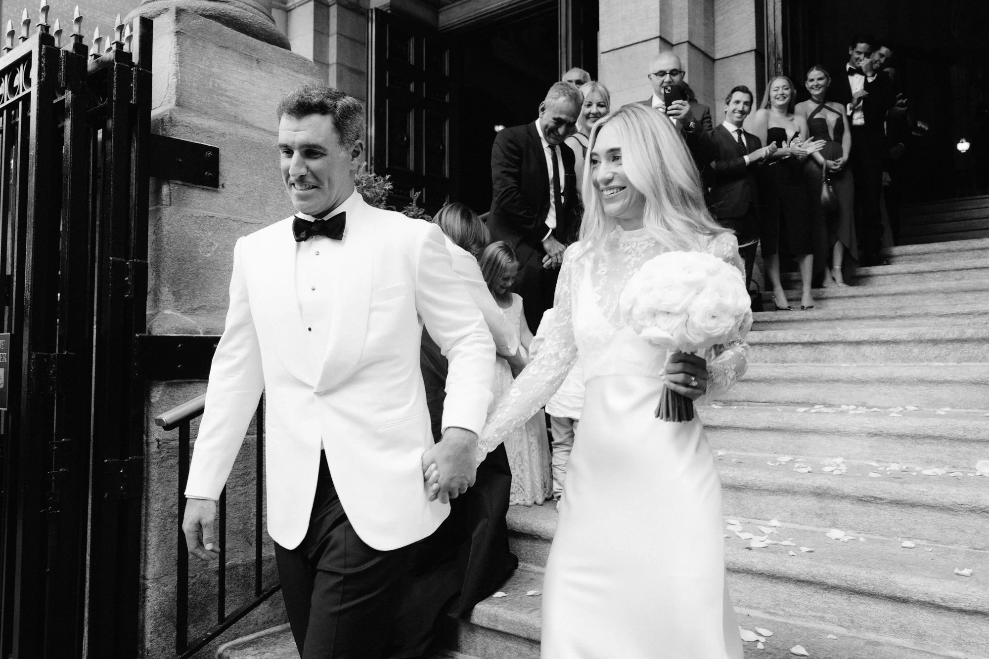 Newlyweds walking hand in hand down the church steps as guests cheer outside St. Francis Xavier Church in Chelsea, photographed by Jenny Fu.