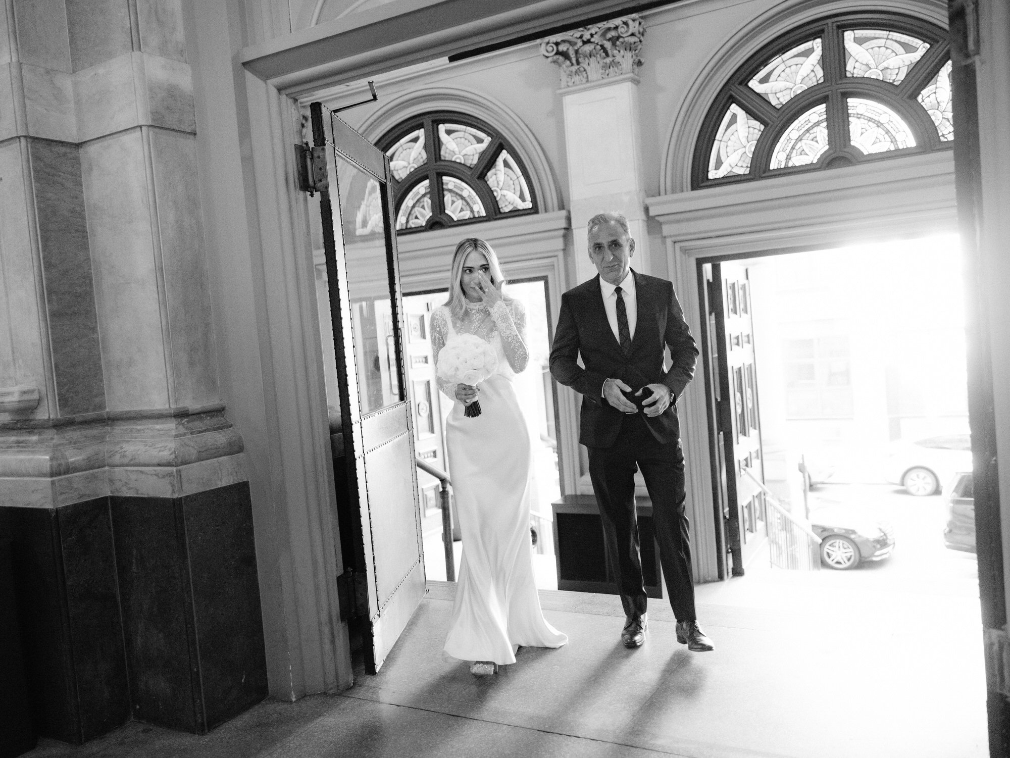 Bride walking into St. Francis Xavier Church on her father’s arm, light streaming through the open doors, photographed by Jenny Fu.