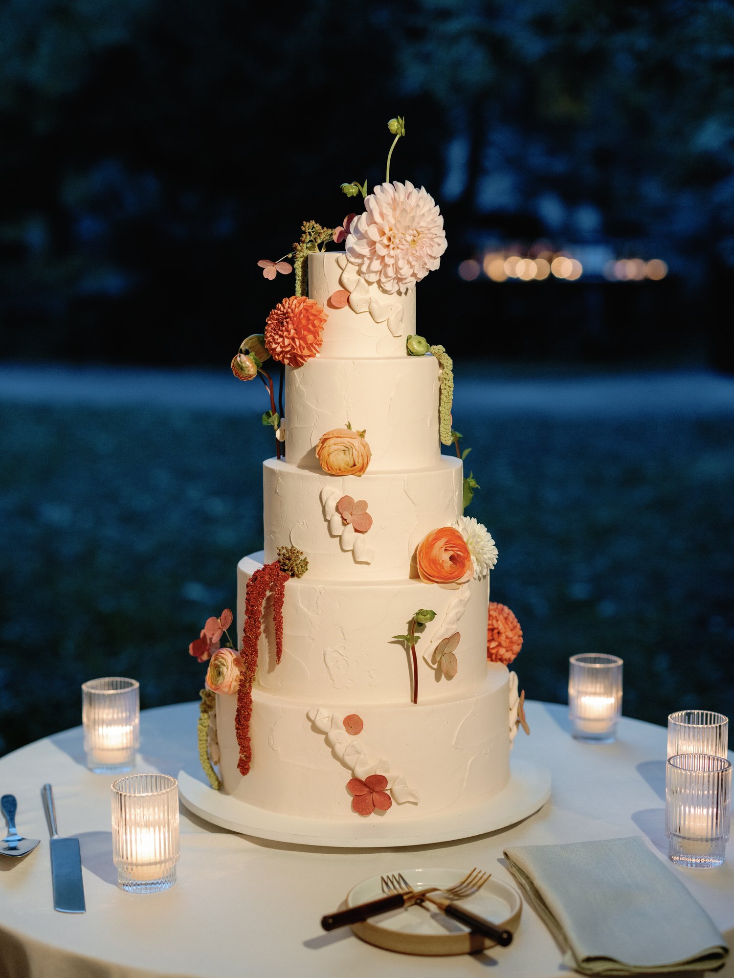 Four tier wedding cake adorned with seasonal flowers on a candlelit table at Troutbeck in Amenia, New York, photographed by Jenny Fu.