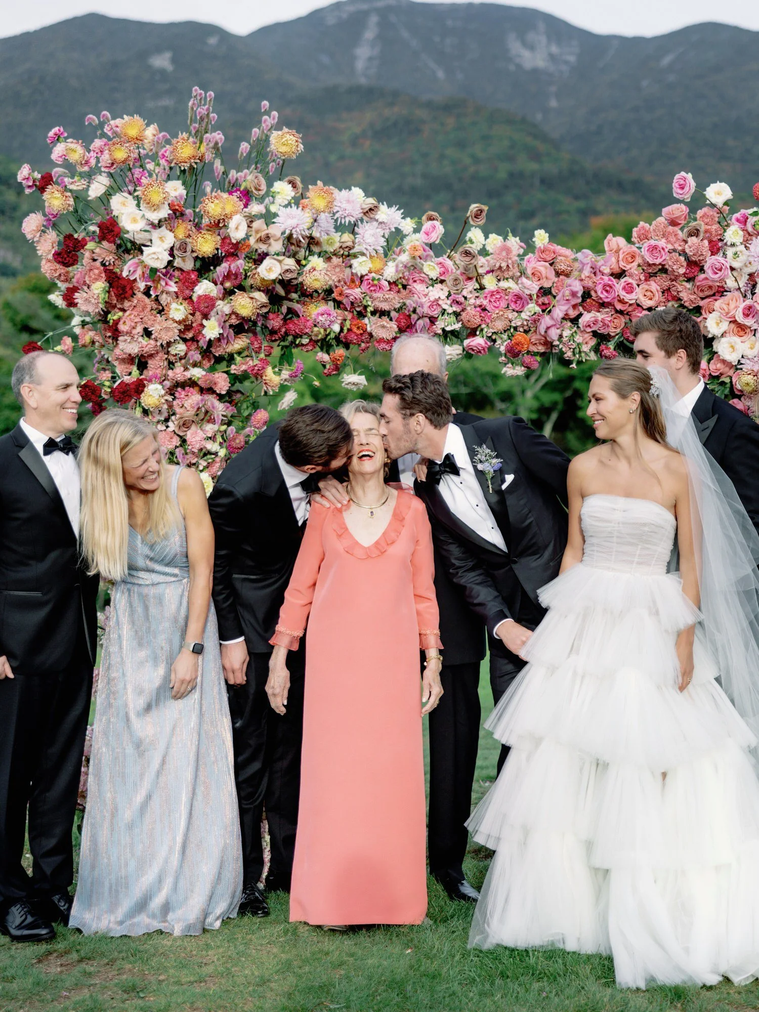 Bride and groom candid moment with family members kissing grandma by floral arch at The Ausable Club in the Adirondacks