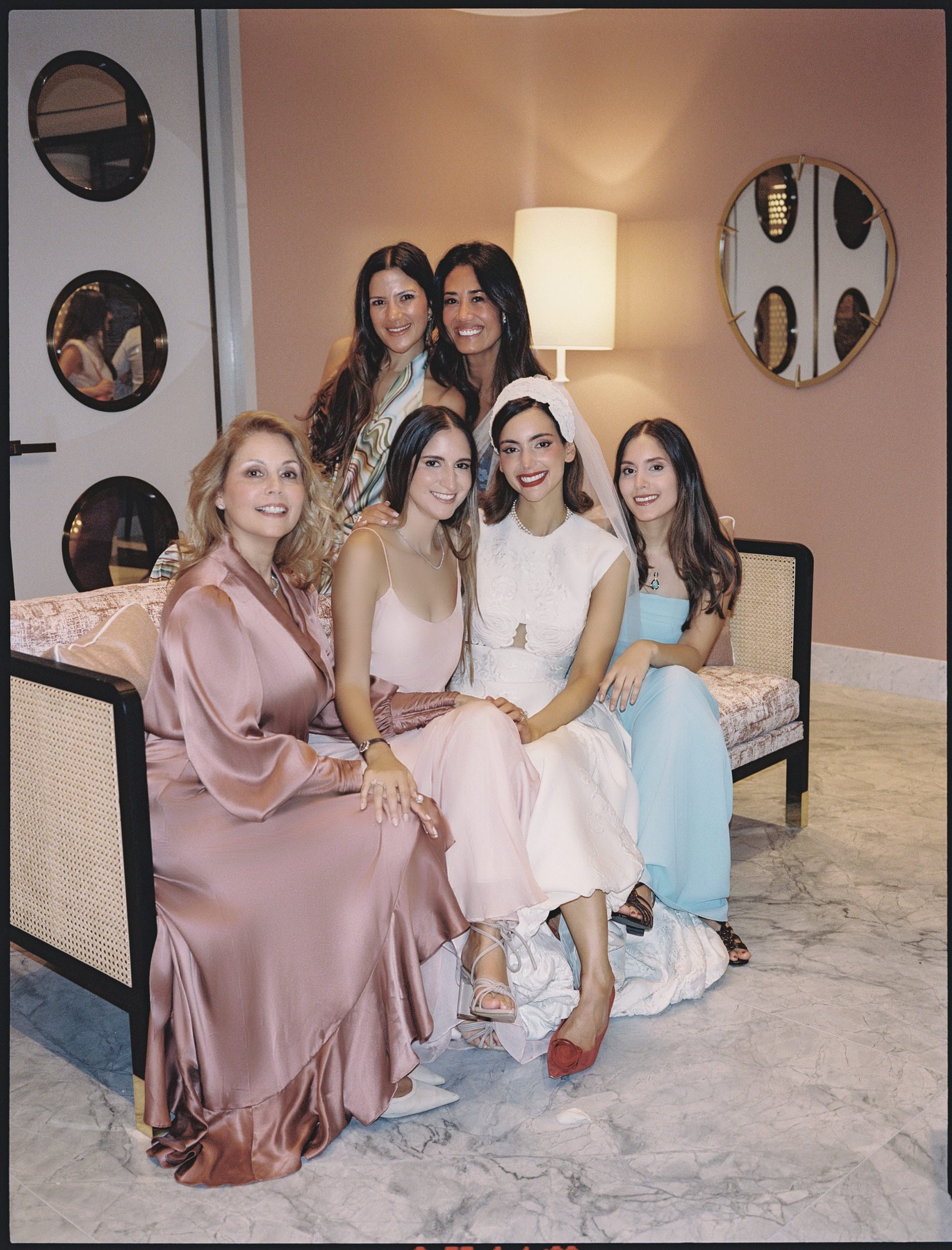 Bride seated with family and bridesmaids in pastel dresses in a blush suite at the Waldorf Astoria New York, photographed by Jenny Fu.