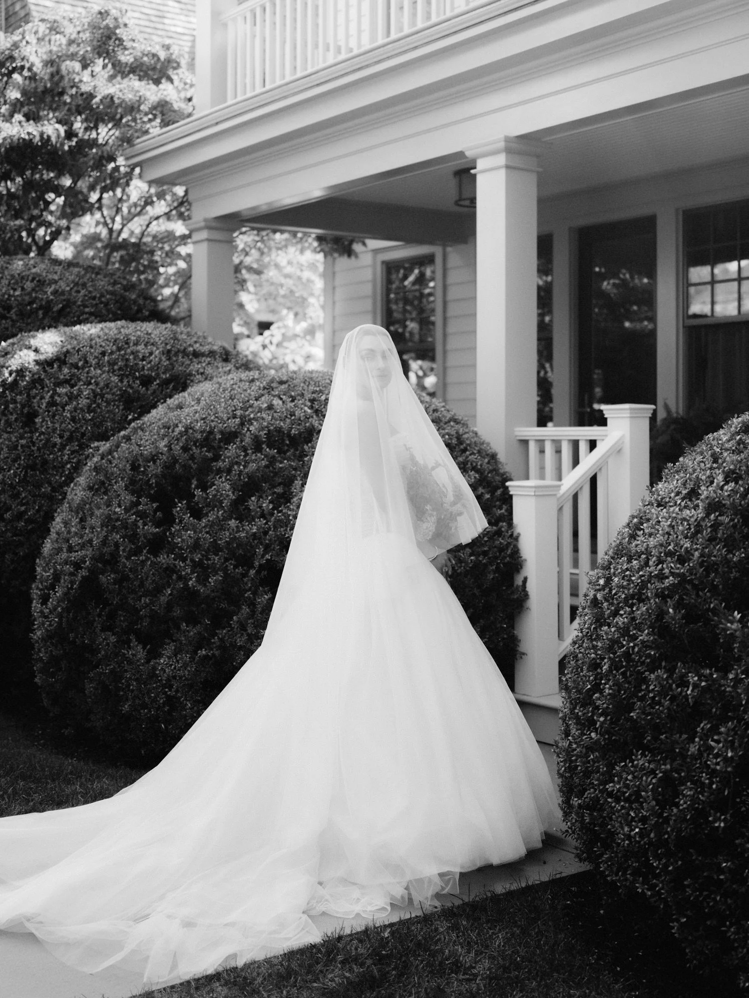 Bride in full tulle gown and long veil looking back beside clipped hedges in a black and white portrait at Valley Rock Inn in Sloatsburg, photographed by Jenny Fu.