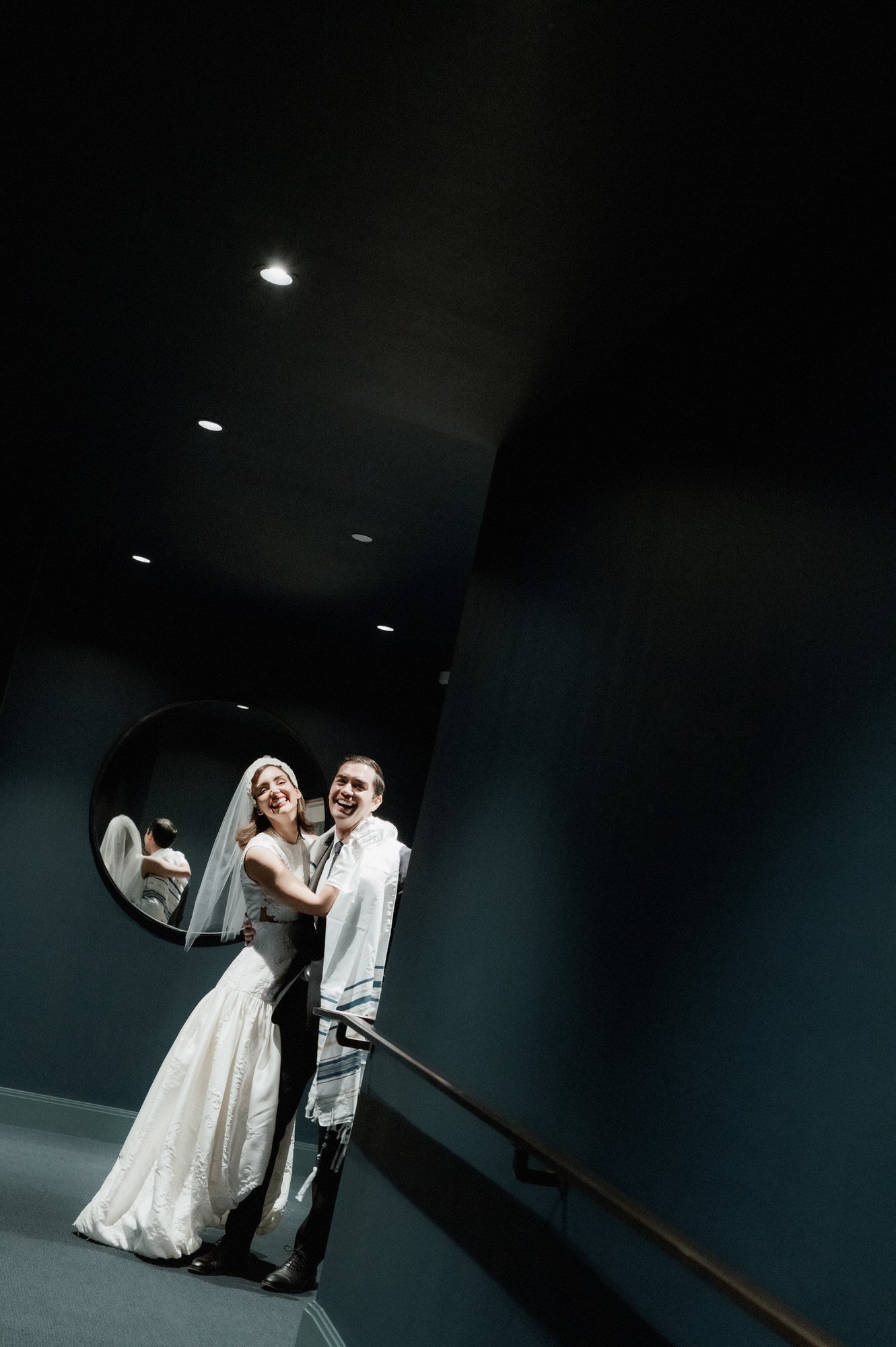 Newlyweds embracing on a dark staircase beneath a round mirror at the Waldorf Astoria New York, photographed by Jenny Fu.