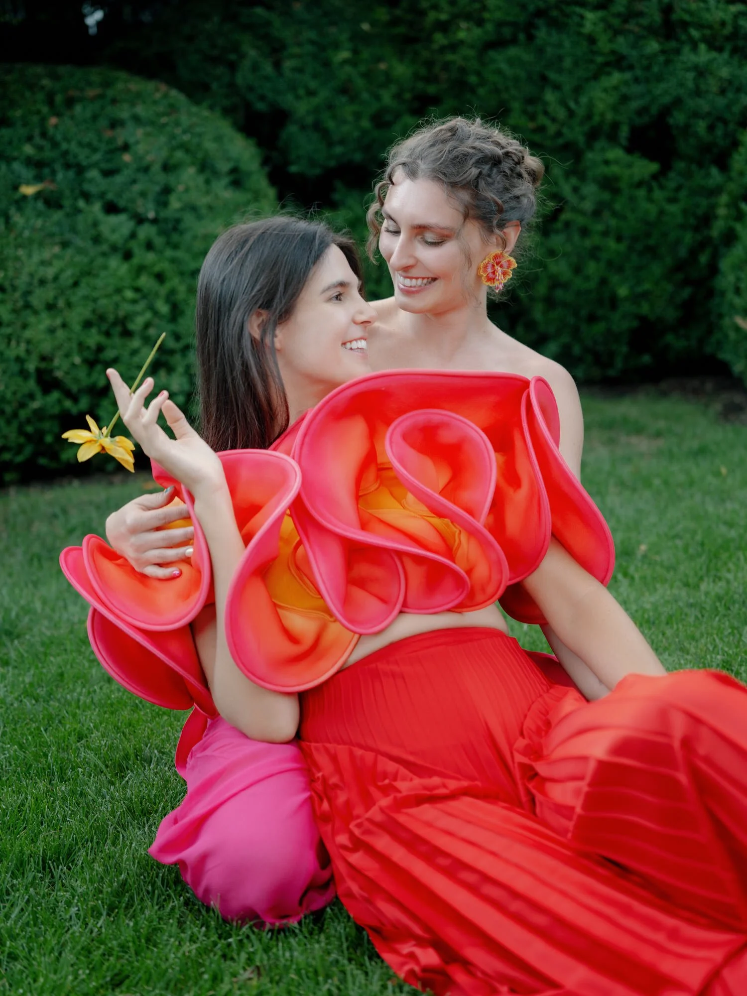 Two brides seated on the lawn, one wearing a sculptural red top, at Valley Rock Inn in Sloatsburg, photographed by Jenny Fu.