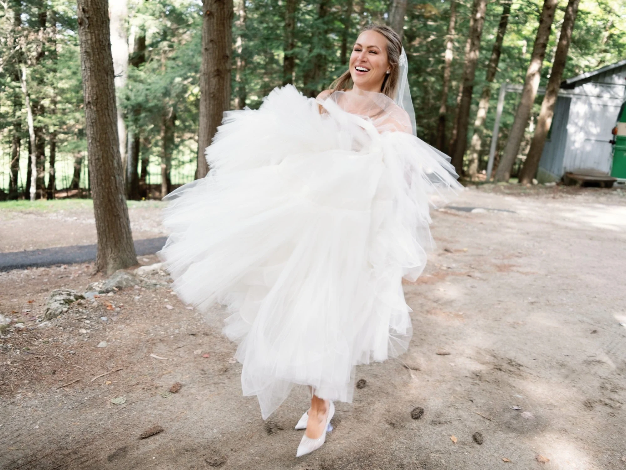 Bride smiling and lifting her tiered gown while walking through wooded path at The Ausable Club in the Adirondacks