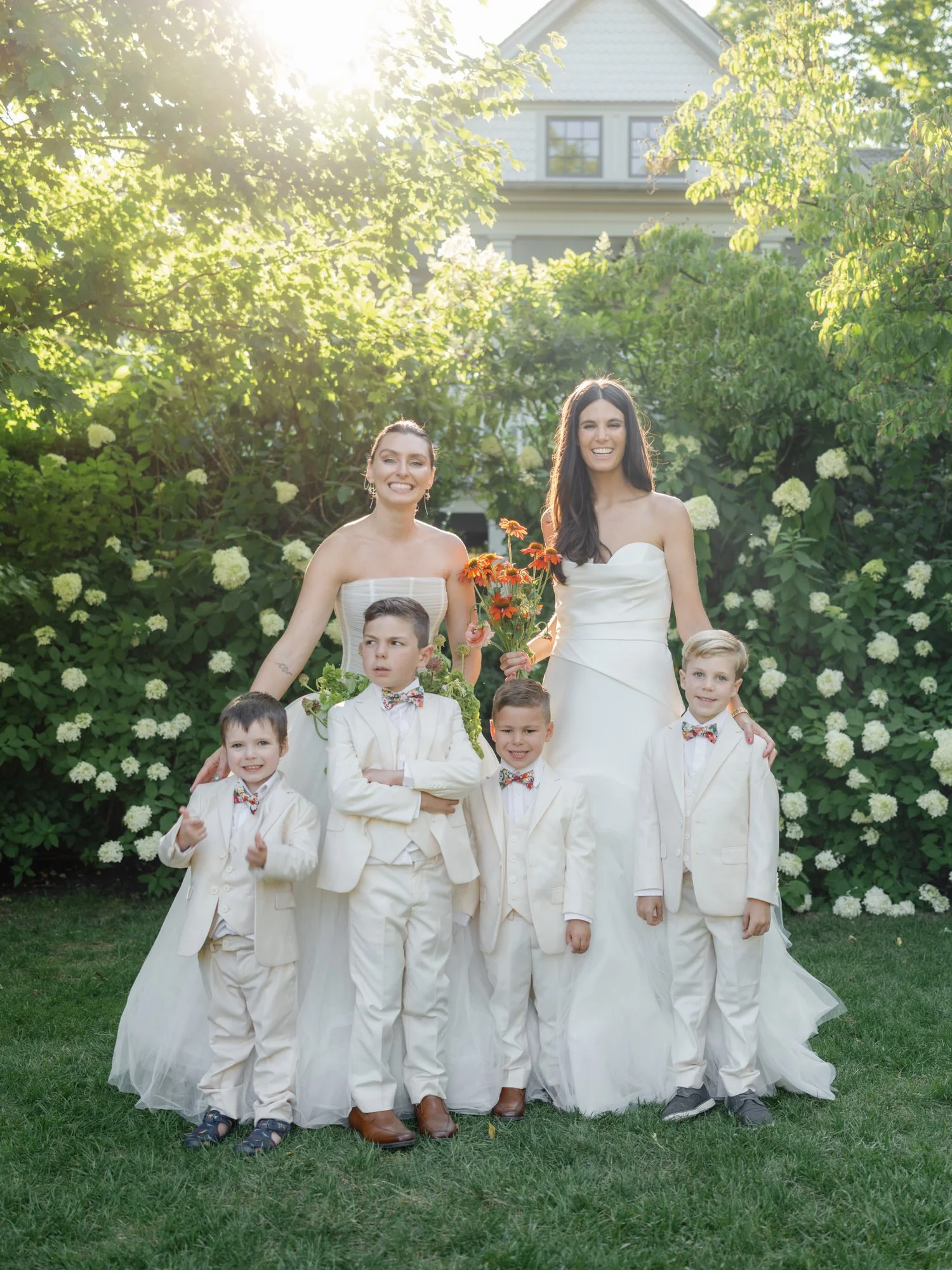 Two brides posing with young ring bearers dressed in cream suits in the garden at Valley Rock Inn in Sloatsburg, photographed by Jenny Fu.