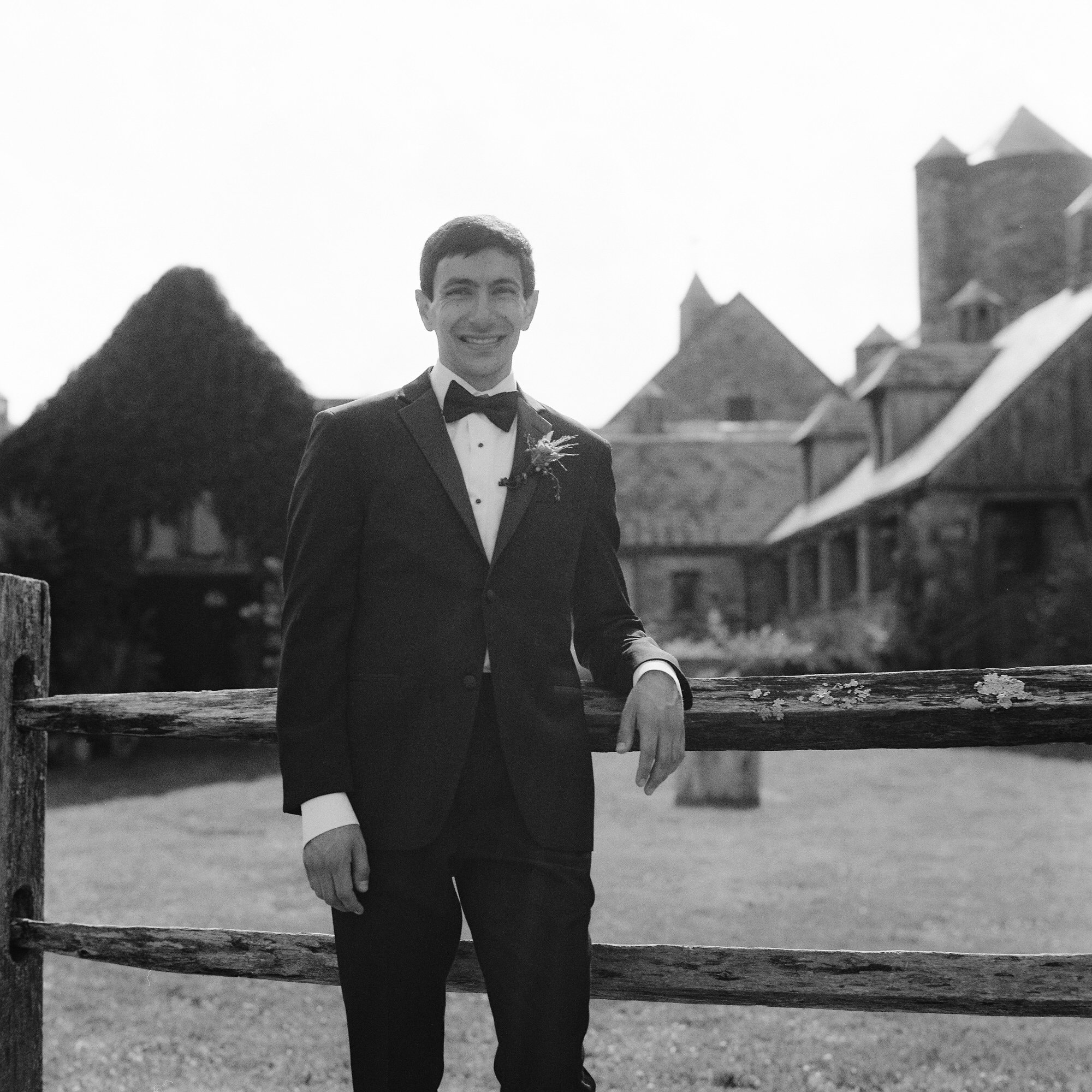 Groom standing by the wooden fence in black and white with the barns in the background at Blue Hill at Stone Barns, photographed by Jenny Fu.