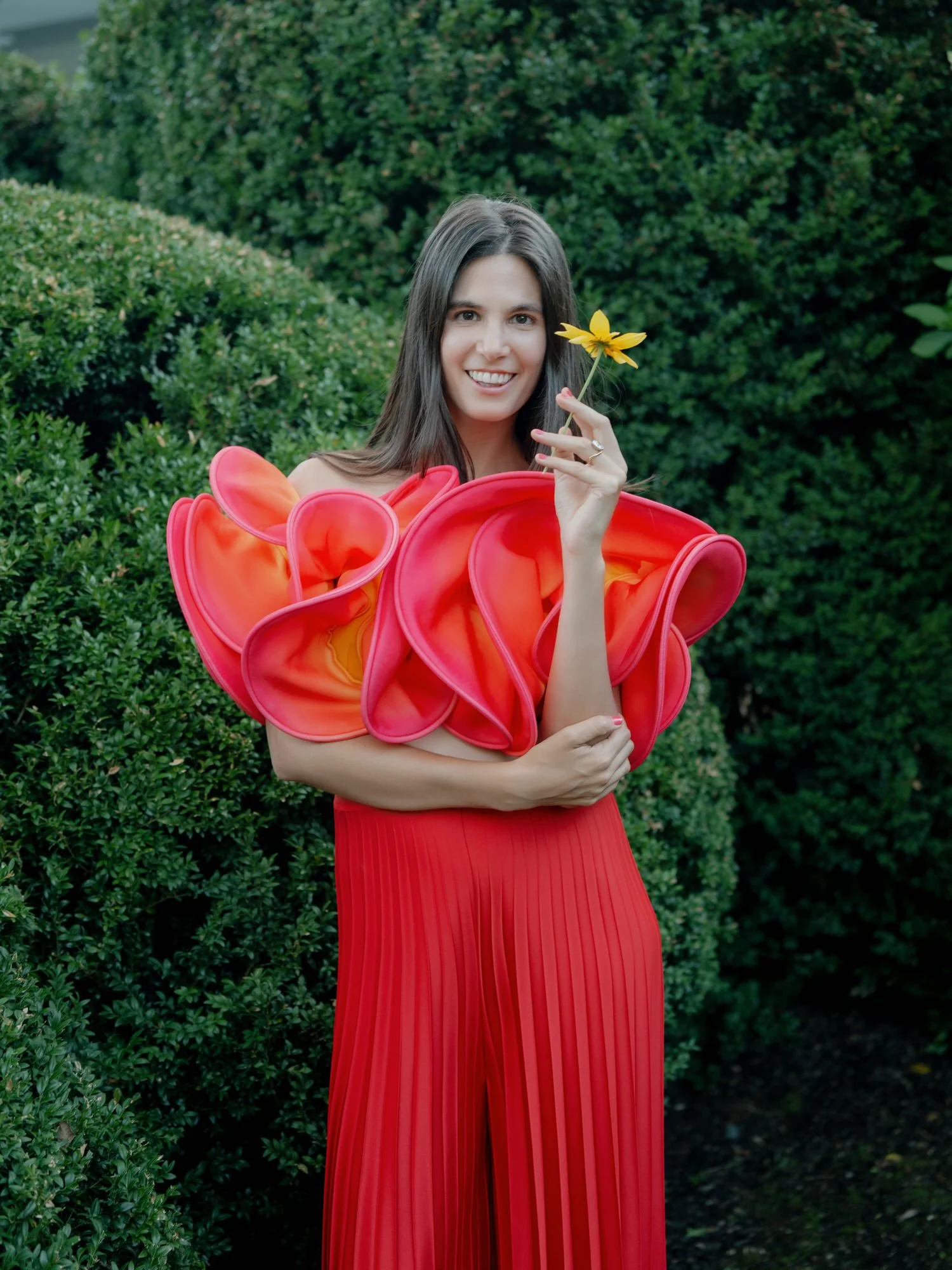 Bride holding a single yellow flower while wearing a sculptural red top in the garden at Valley Rock Inn in Sloatsburg, photographed by Jenny Fu.