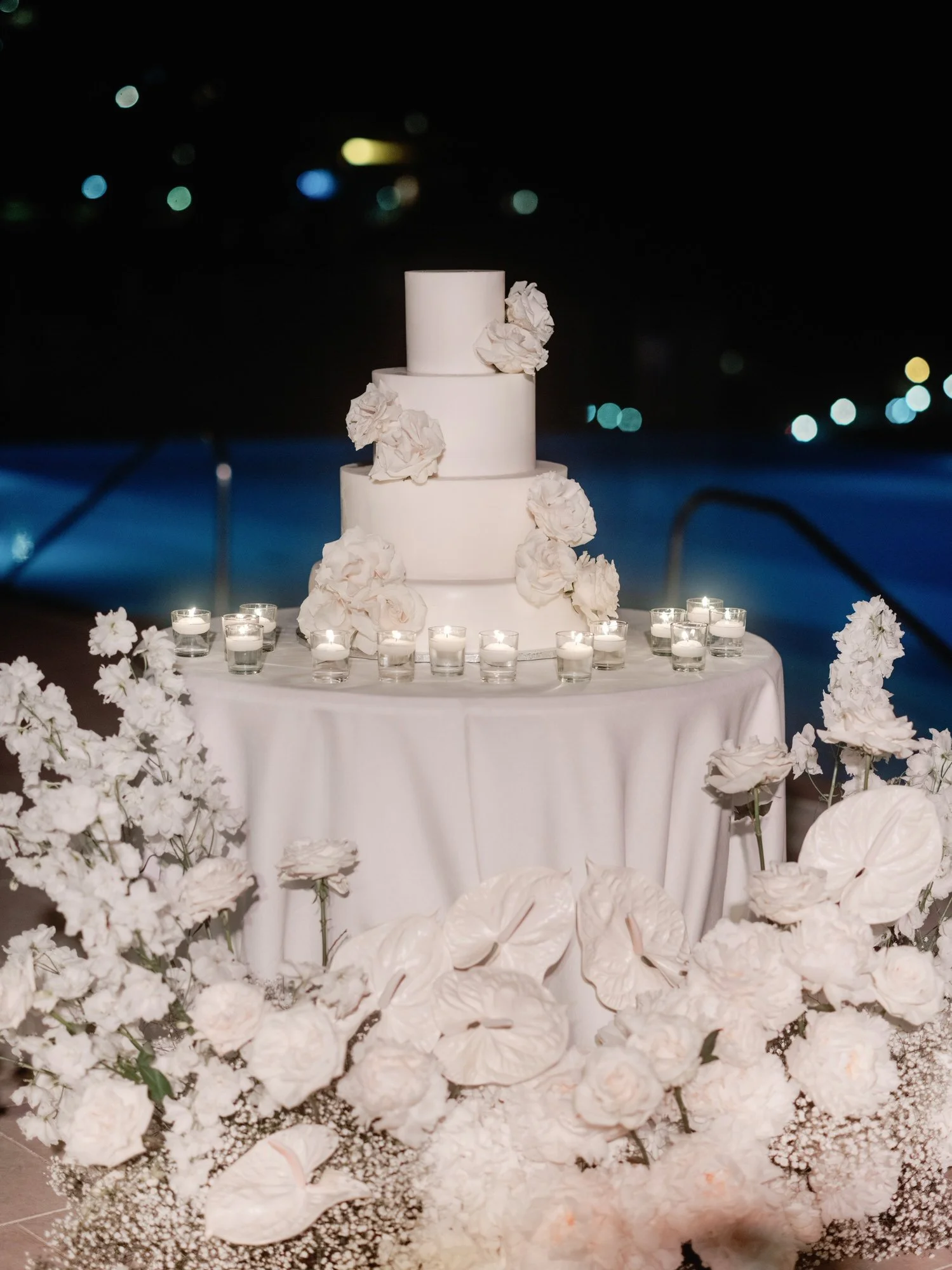 Three-tier white wedding cake adorned with florals and surrounded by candles by the pool at Caruso, A Belmond Hotel in Ravello, Italy, photographed by Jenny Fu.