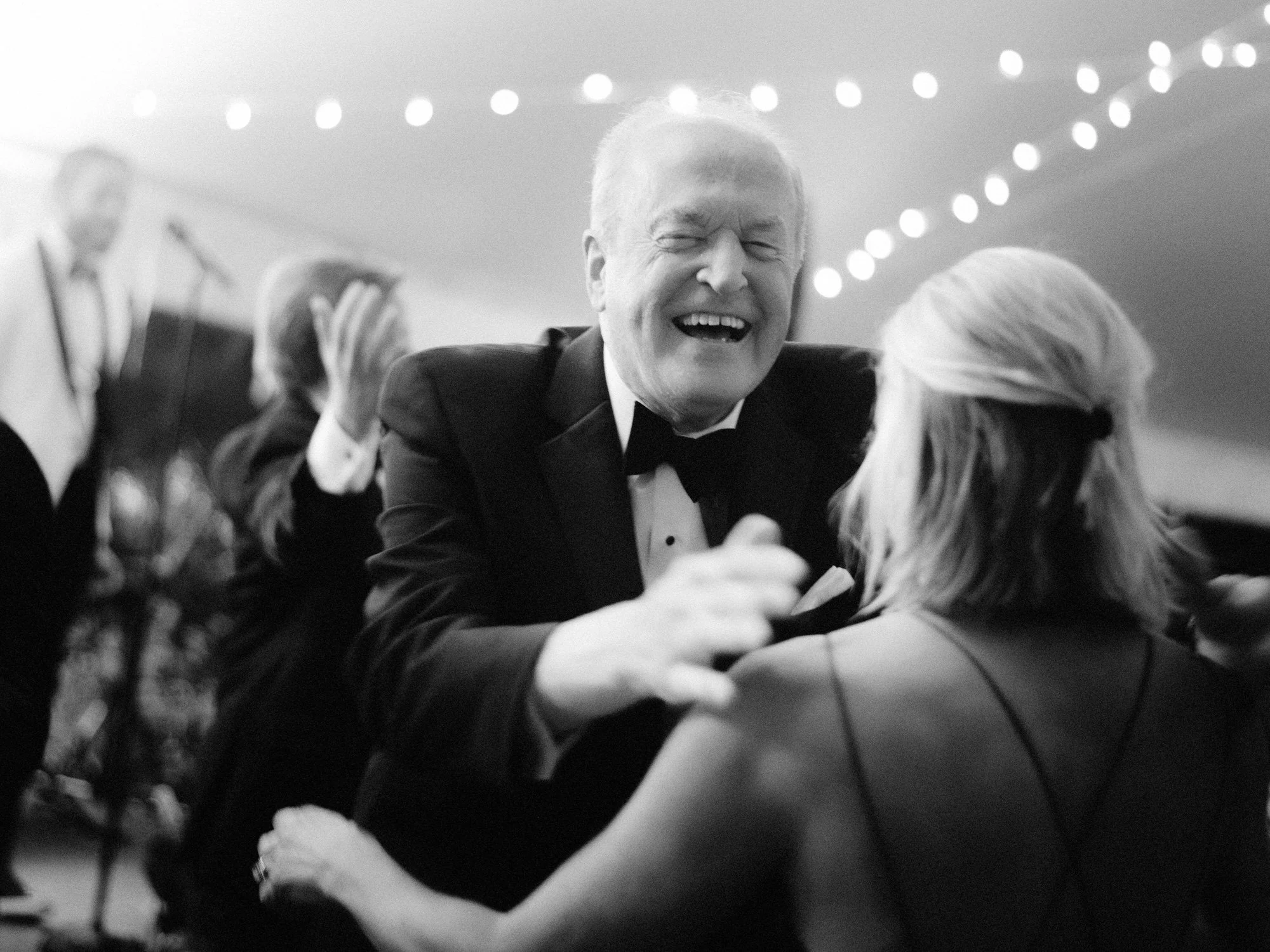 A tuxedoed grandpa bursts into laughter while dancing beneath string lights at the Ausable Club in the Adirondacks
