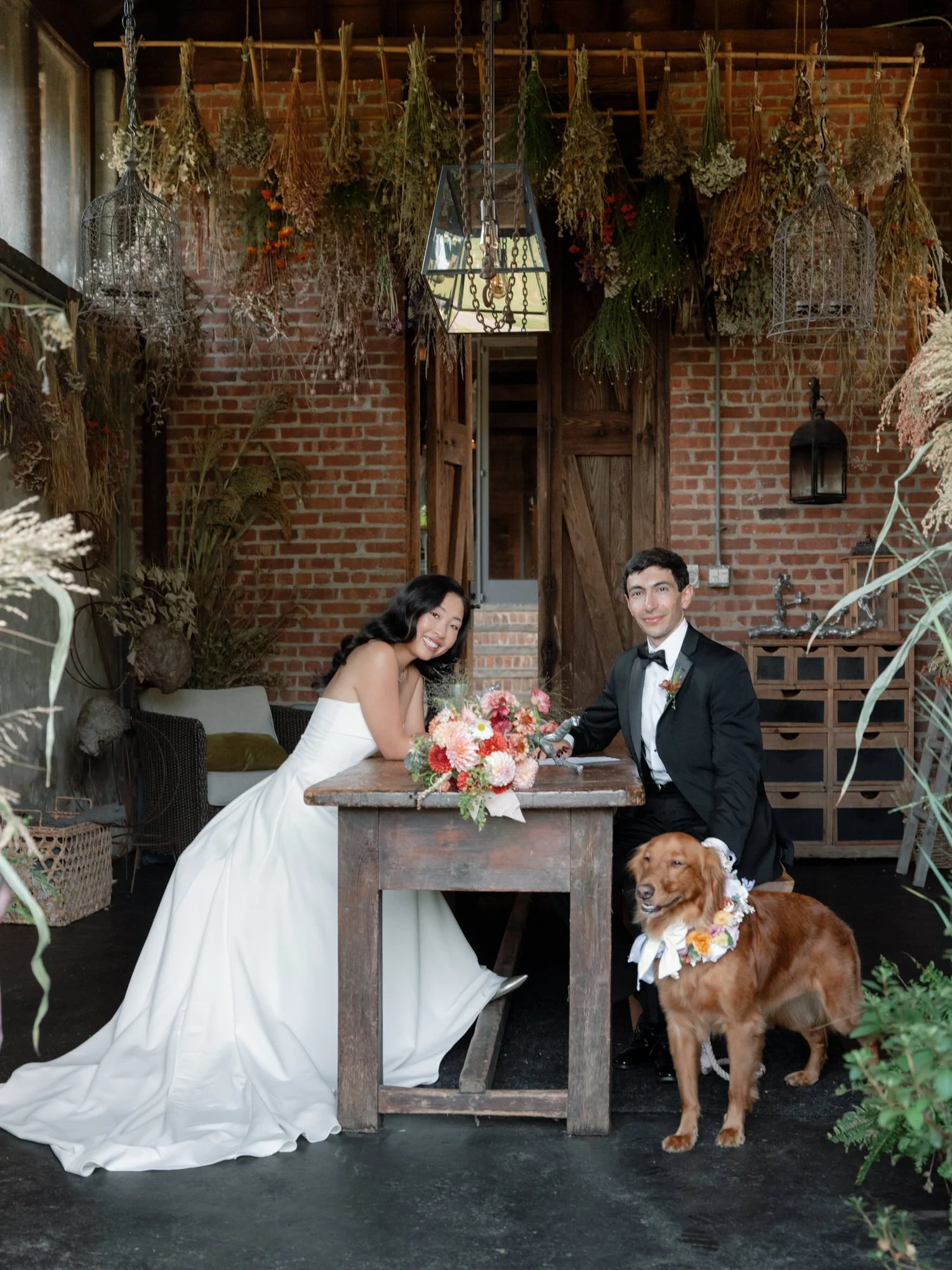 Bride and groom seated at a rustic table in the drying room with their flower-collared dog beside them at Blue Hill at Stone Barns, photographed by Jenny Fu.